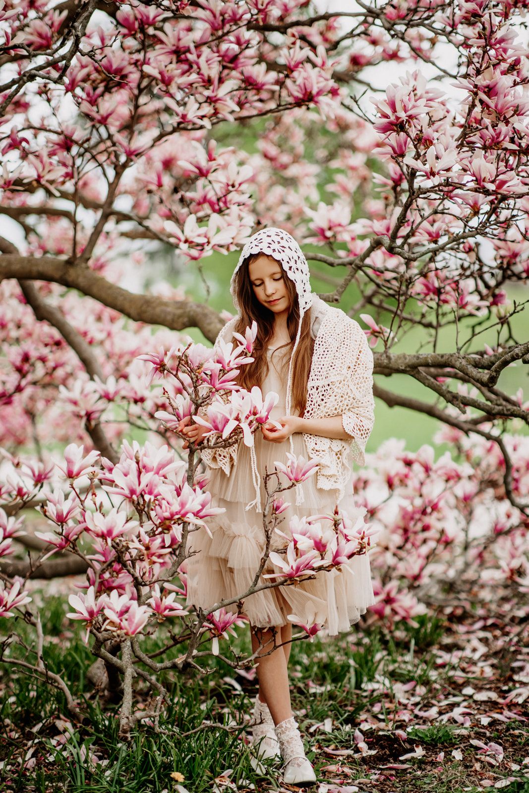 Young woman in lace hood and tiered dress admiring pink magnolia blossoms in a spring garden.