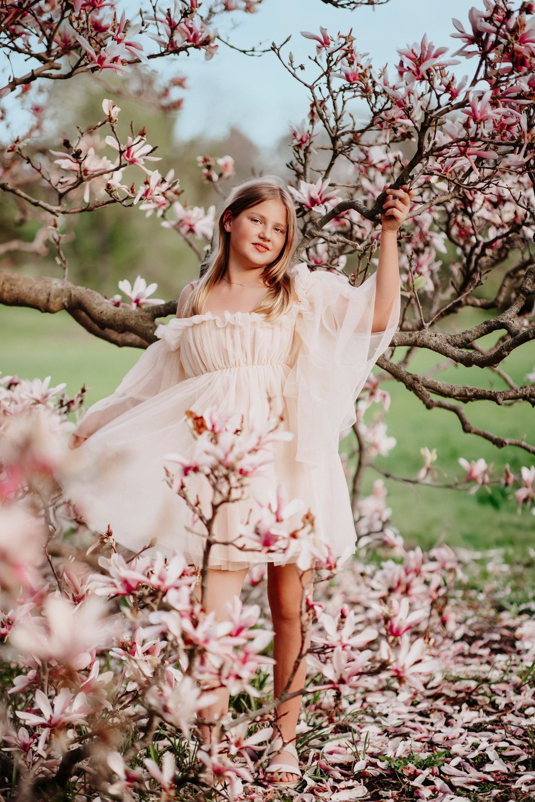 Young girl in flowing white dress posing among blooming magnolia trees outdoors.