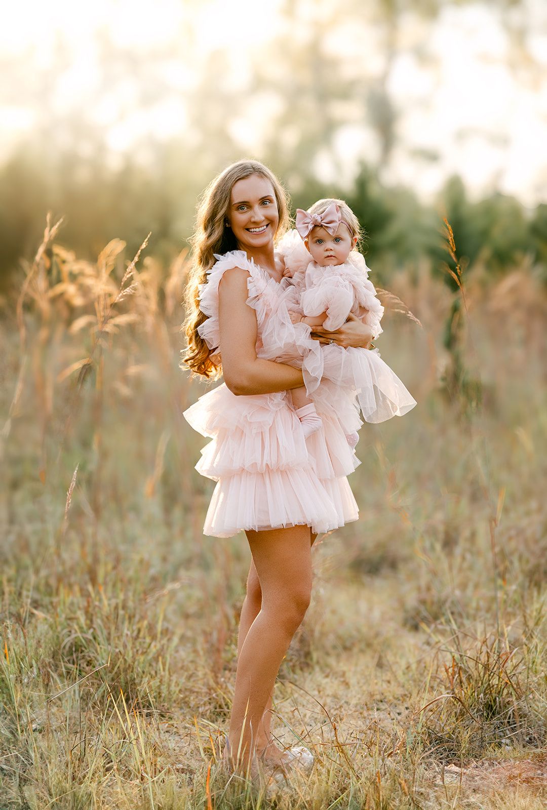 Mom and baby girl in matching pink tiered ruffle dresses pose in sunlit meadow at golden hour.