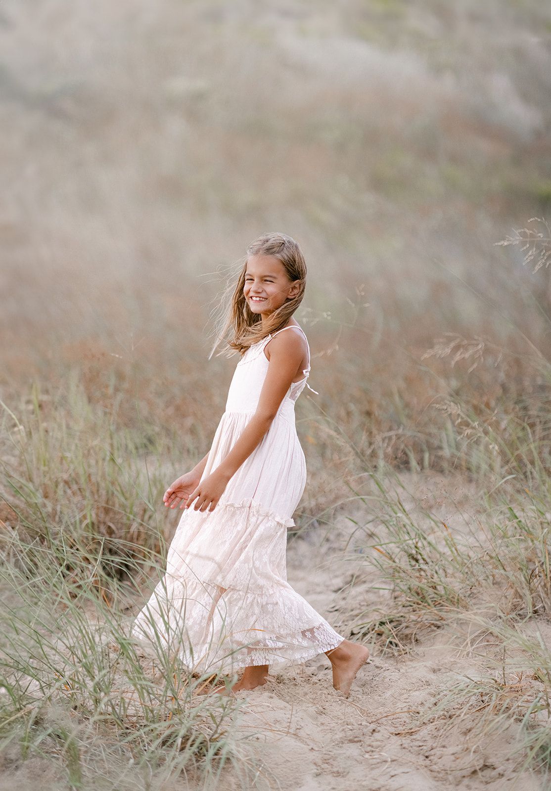 Young girl in white lace dress walking barefoot through sandy beach dunes, smiling joyfully.