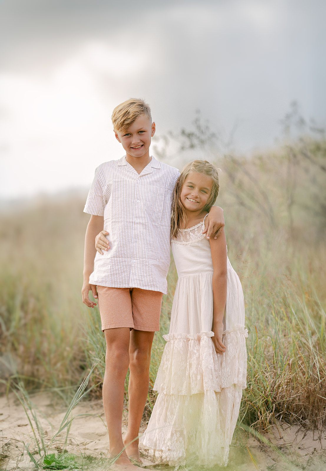 Two smiling children pose together on sandy dunes, boy in white shirt and pink shorts, girl in white lace dress.