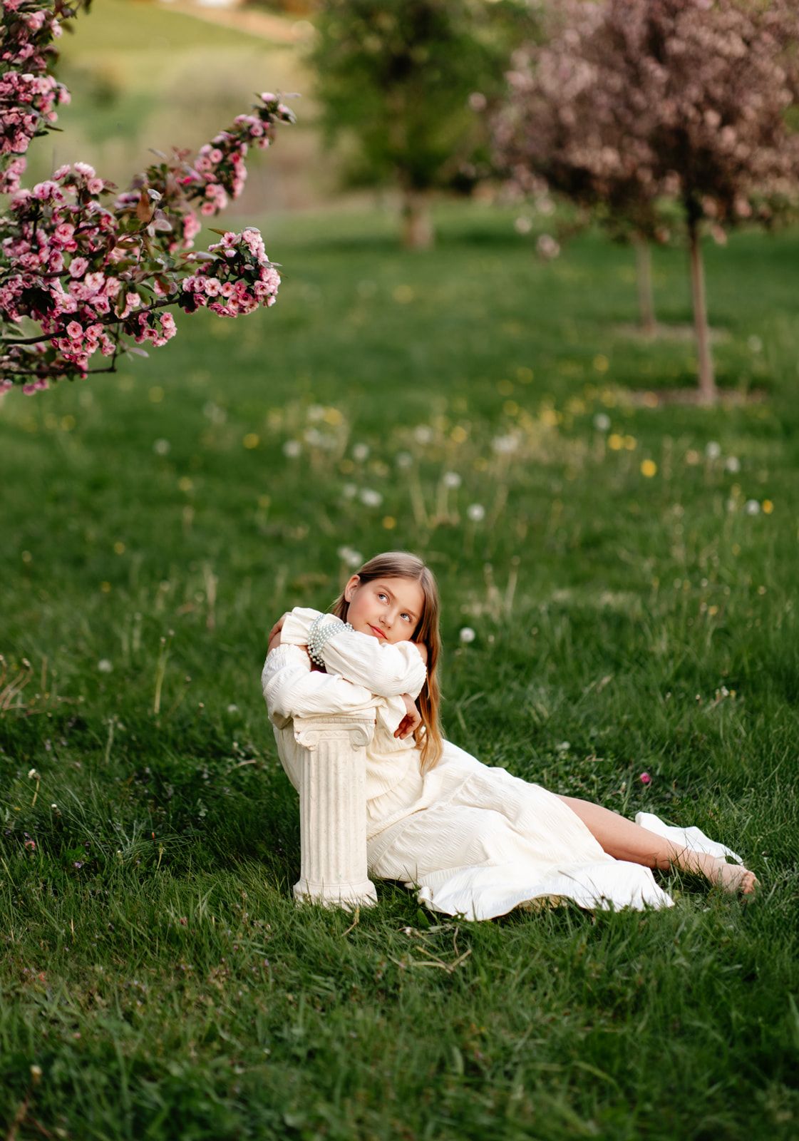 Young girl in cream dress sits on grass leaning on white pillar near pink blossoming trees outdoors.