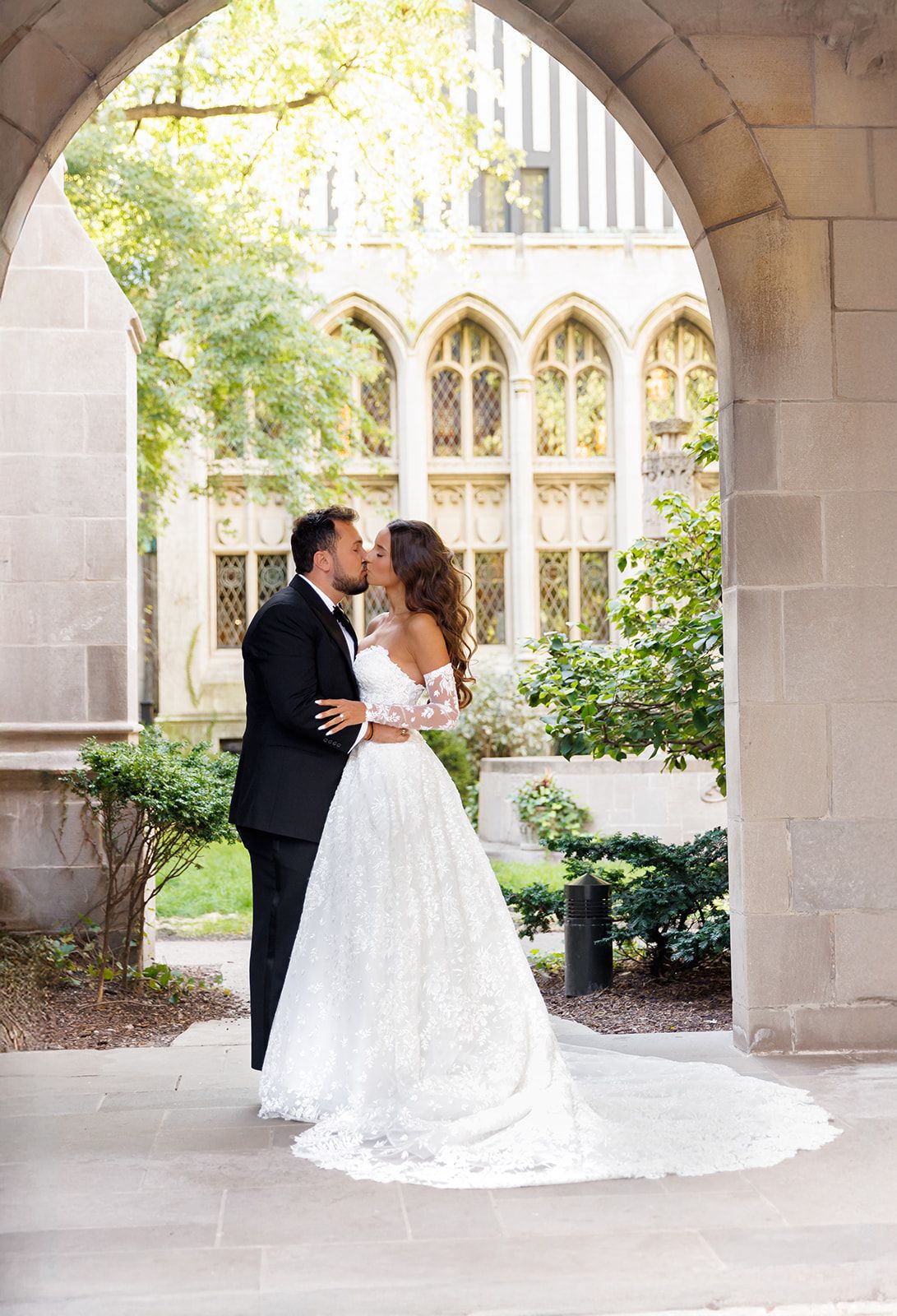 Newlyweds kiss under a stone arch with lush greenery, bride in an off-shoulder lace gown with a long train.