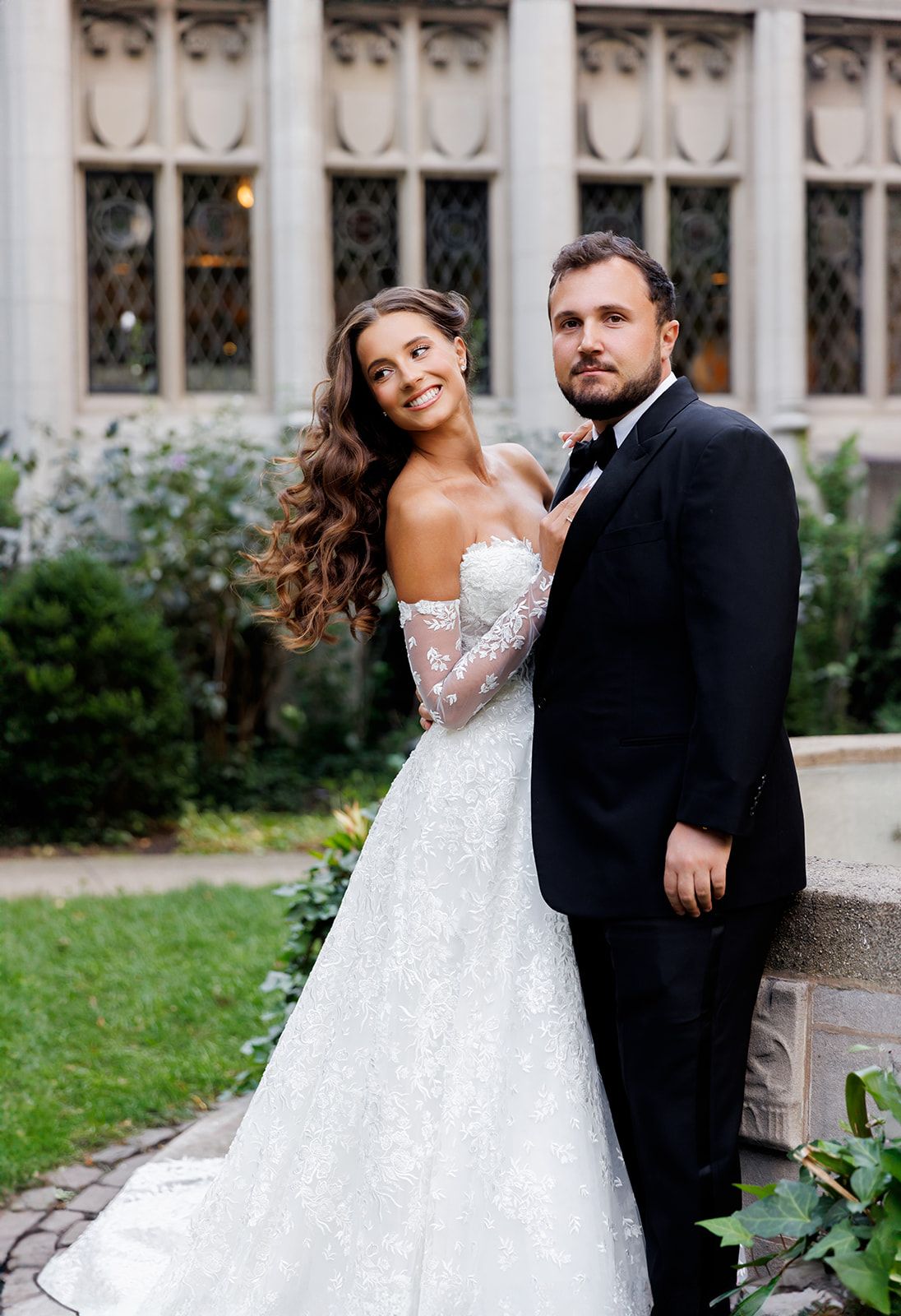 Bride in lace off-shoulder gown with groom in black tuxedo posing outdoors at elegant wedding venue.