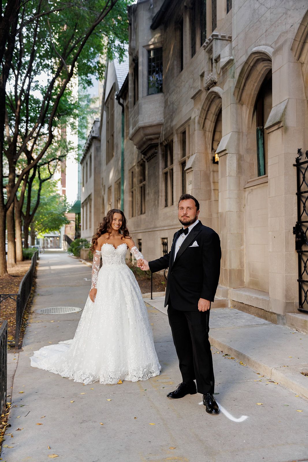 Bride in lace gown and groom in tuxedo hold hands on urban sidewalk beside historic stone building.