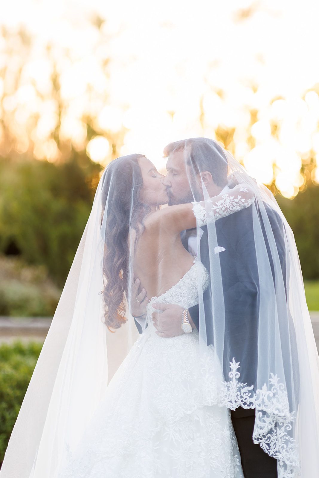 Wedding couple kissing tenderly under a lace-trimmed veil at golden hour in a garden setting.