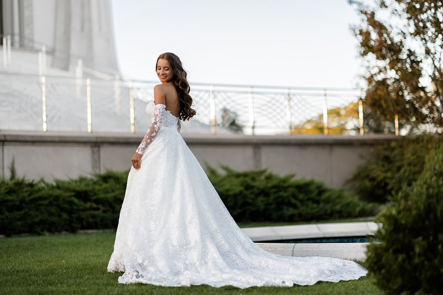 Bride in off-shoulder lace wedding gown with long train poses gracefully outdoors near manicured gardens.