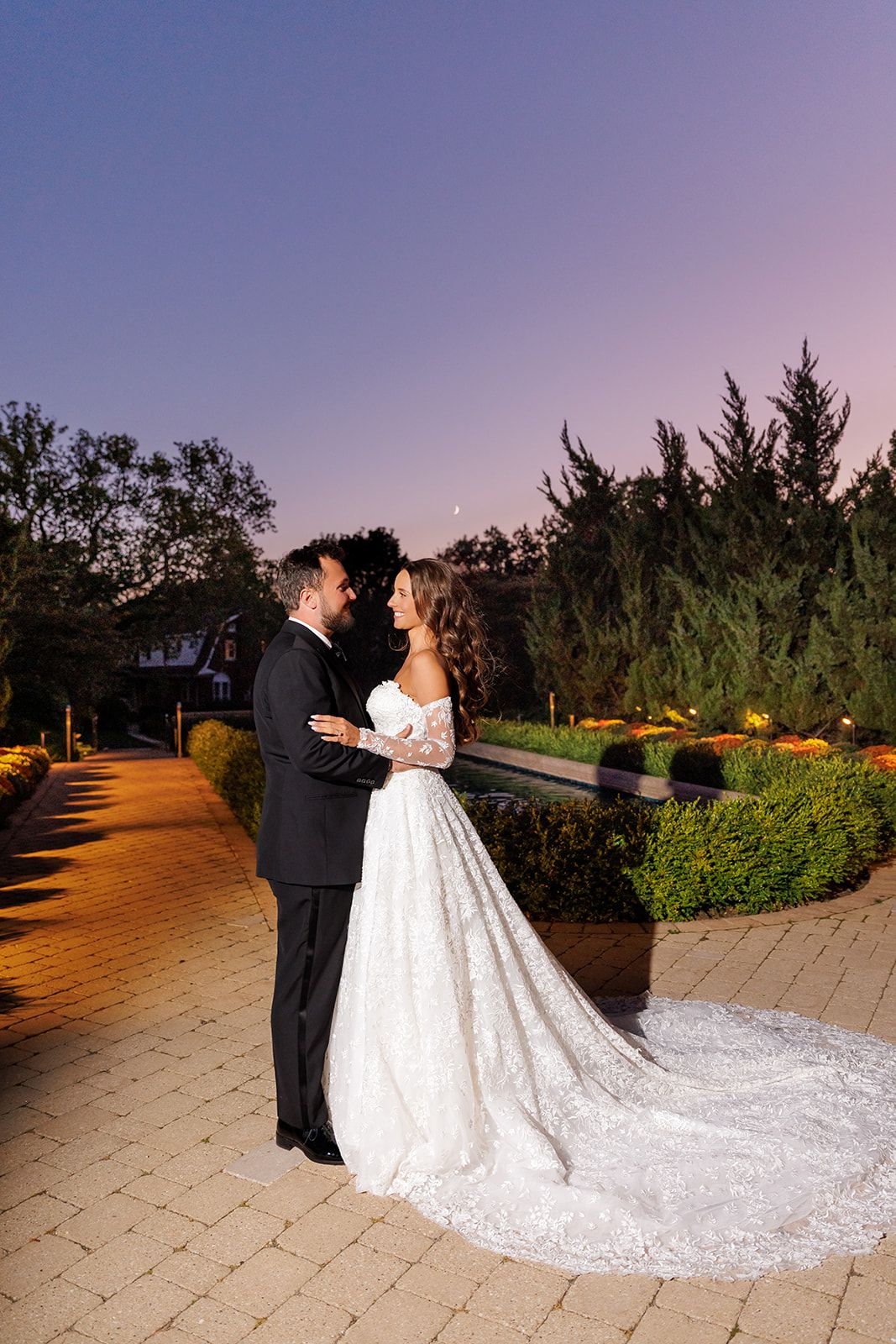 Bride in white lace gown and groom in black tuxedo embrace on a garden path at twilight.