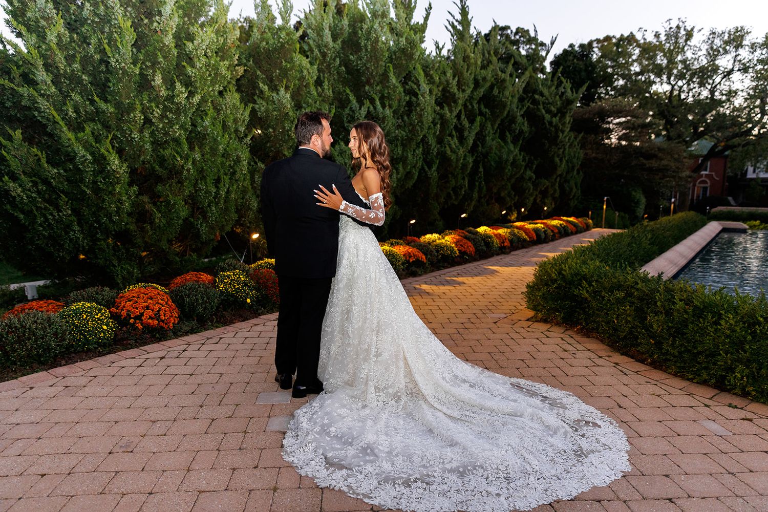 Bride and groom share romantic embrace in garden with colorful chrysanthemums and evergreen trees at dusk.