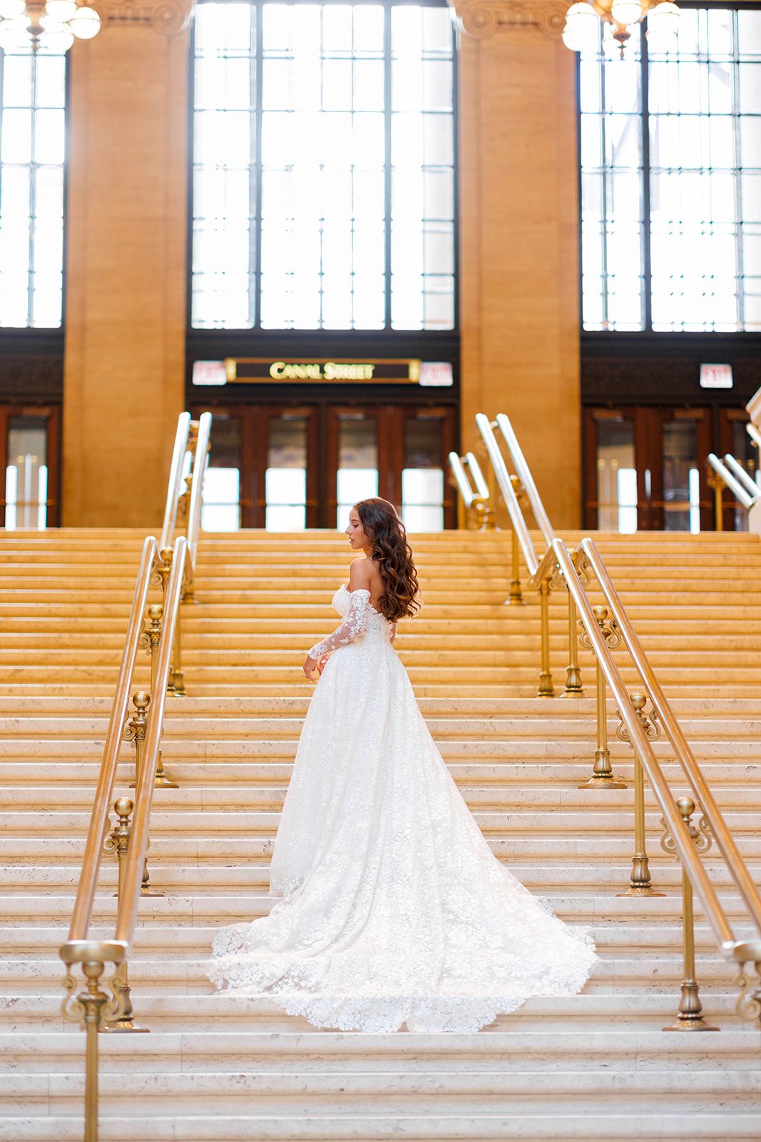 Bride in a white lace gown with long train stands on grand staircase at Canal Street Station.