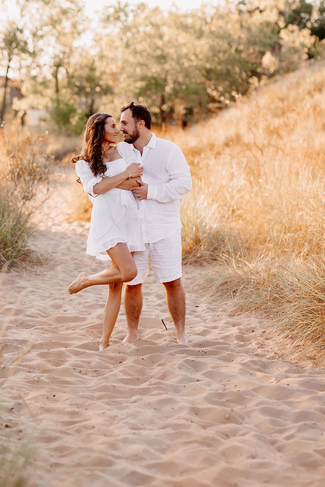 Man lifting woman in white dress while embracing on sandy path with golden grass.