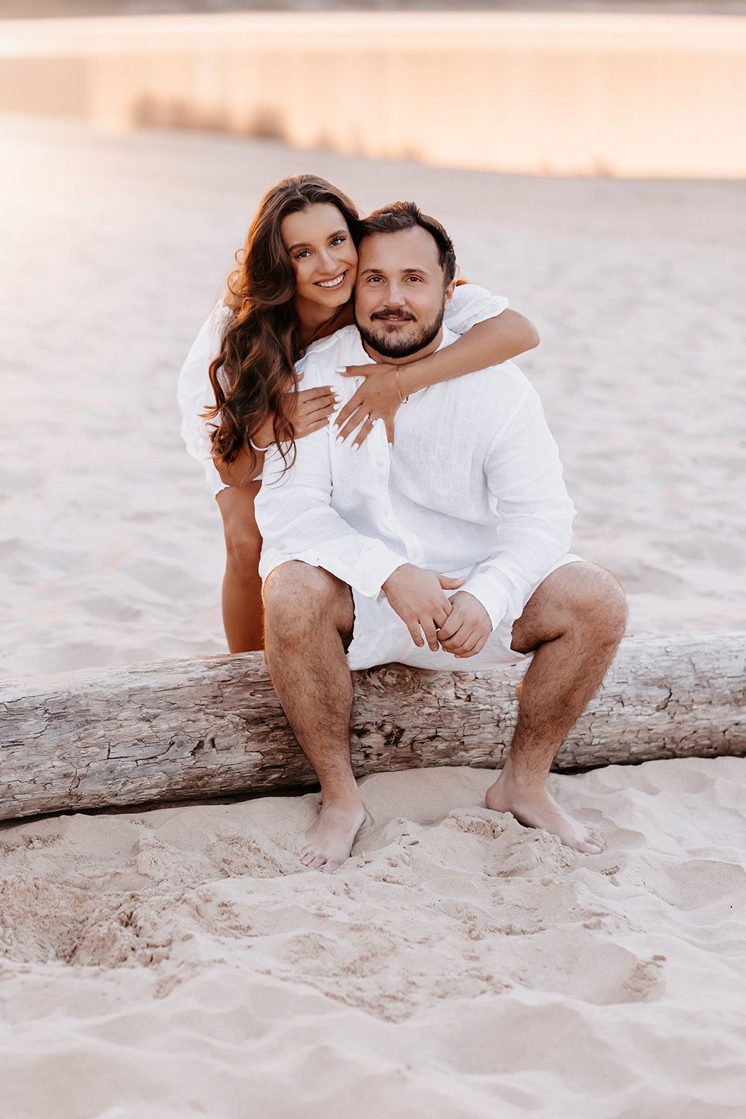 Couple in white outfits sitting on driftwood on sandy beach, woman hugging man from behind at sunset.