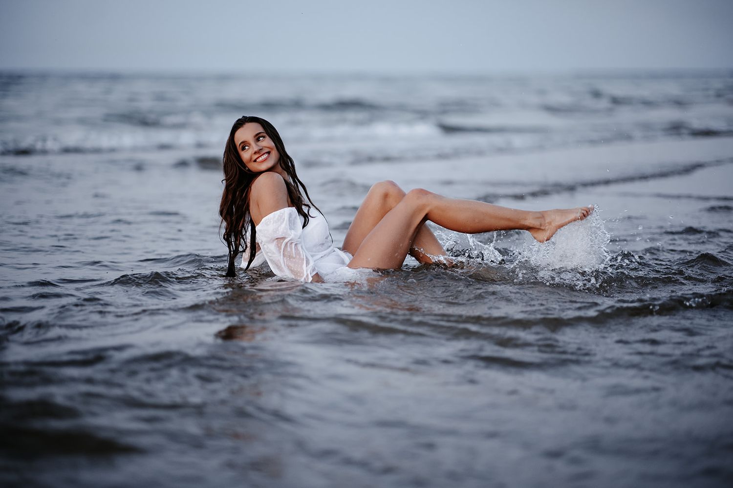 A woman in a white off-shoulder dress sits in shallow ocean waves, smiling with wet hair on a cloudy day.