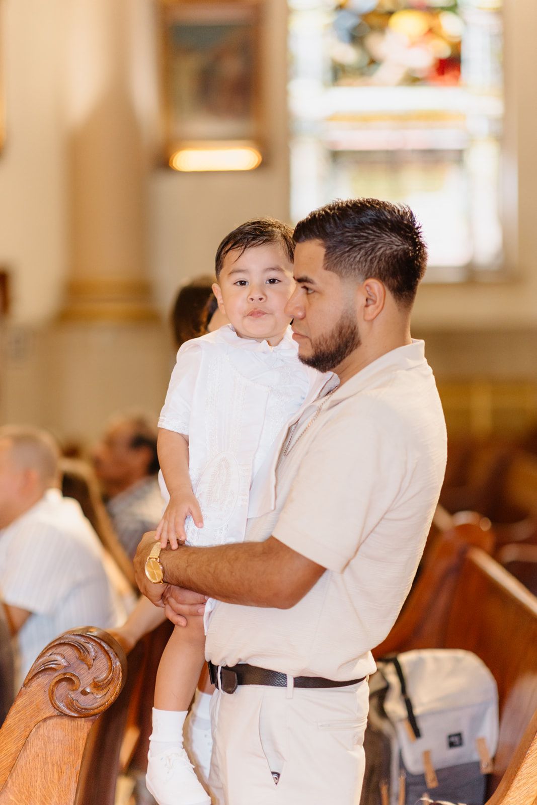 A father holds his young son dressed in white during a church baptism ceremony, sharing a tender kiss on the cheek.