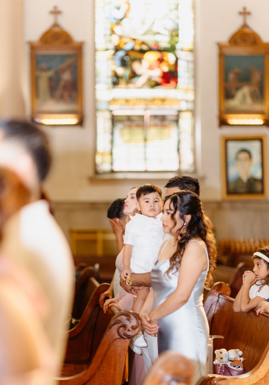 Bride in white gown holds young child during church wedding ceremony with stained glass windows.