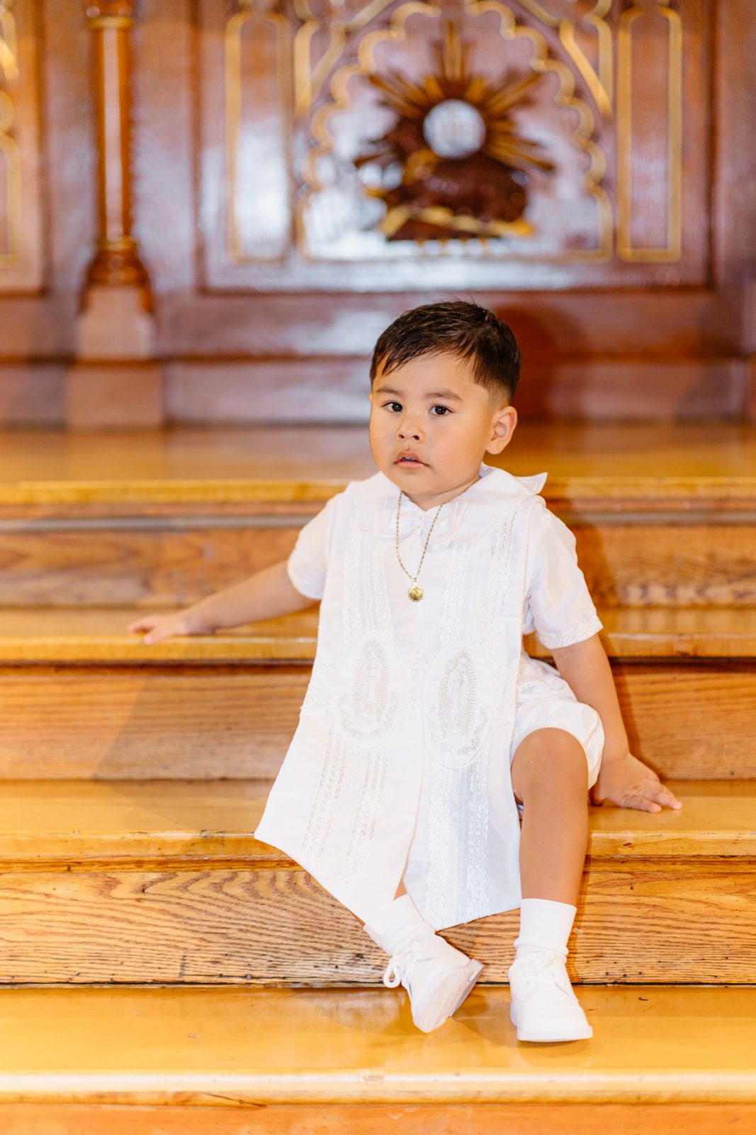 Toddler boy in white barong outfit sitting on wooden church steps during baptism ceremony.