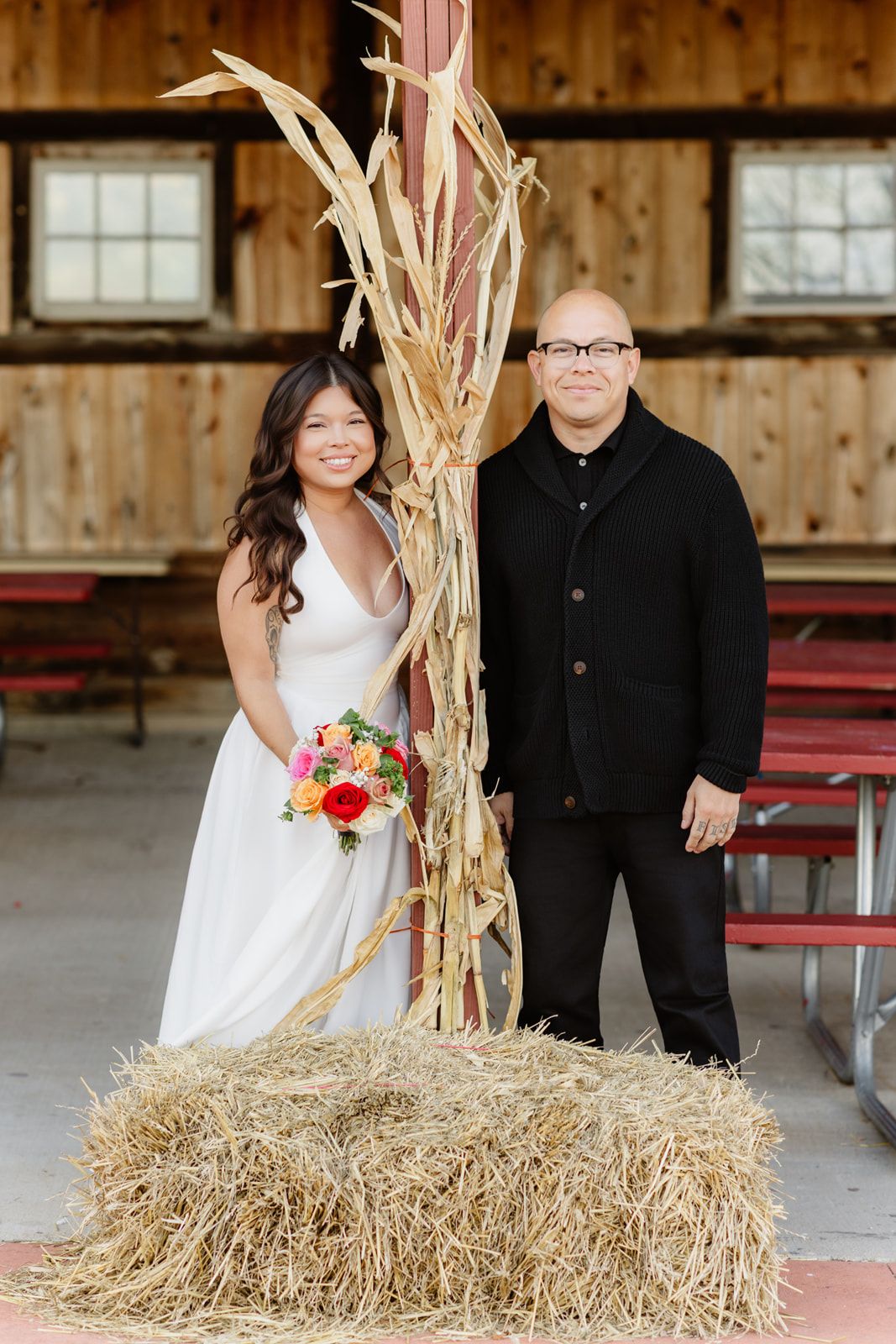 Bride in white gown holds colorful bouquet beside groom in black, posing near hay bale in rustic barn setting.
