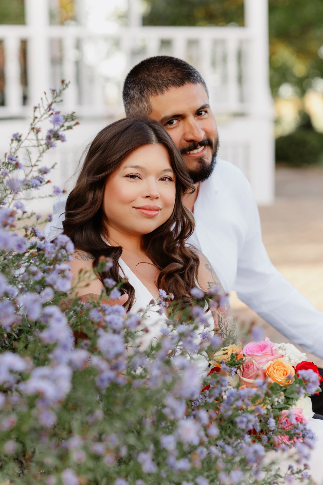 Couple smiling among purple wildflowers, woman holding colorful bouquet, man in white shirt, romantic outdoor portrait.