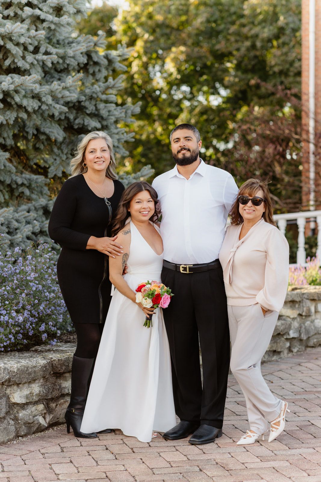 Bride in white dress holding bouquet poses with groom and two women guests on brick patio at outdoor wedding.