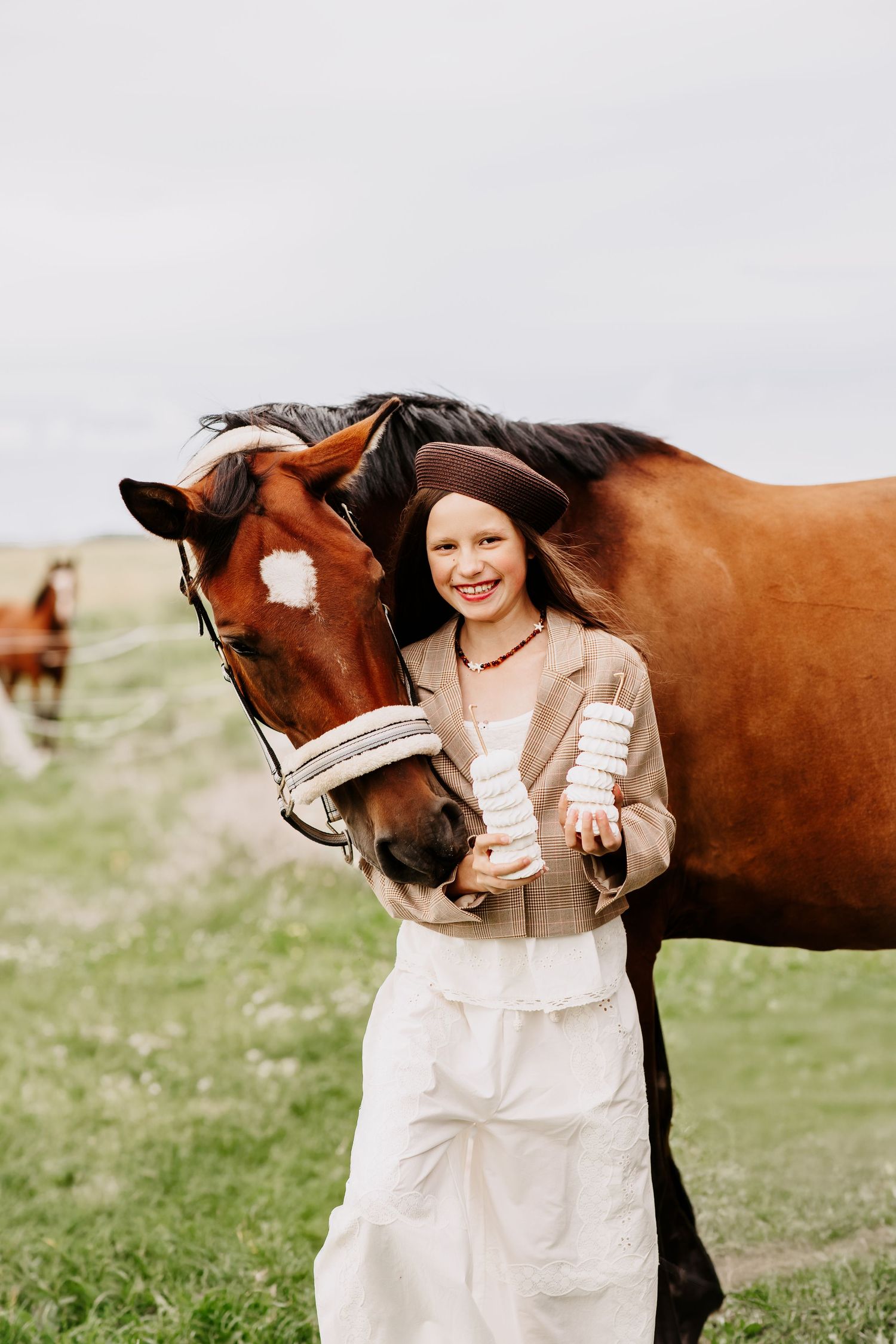 Smiling girl in brown beret and beige blazer posing with a chestnut horse while holding white treat stacks outdoors.
