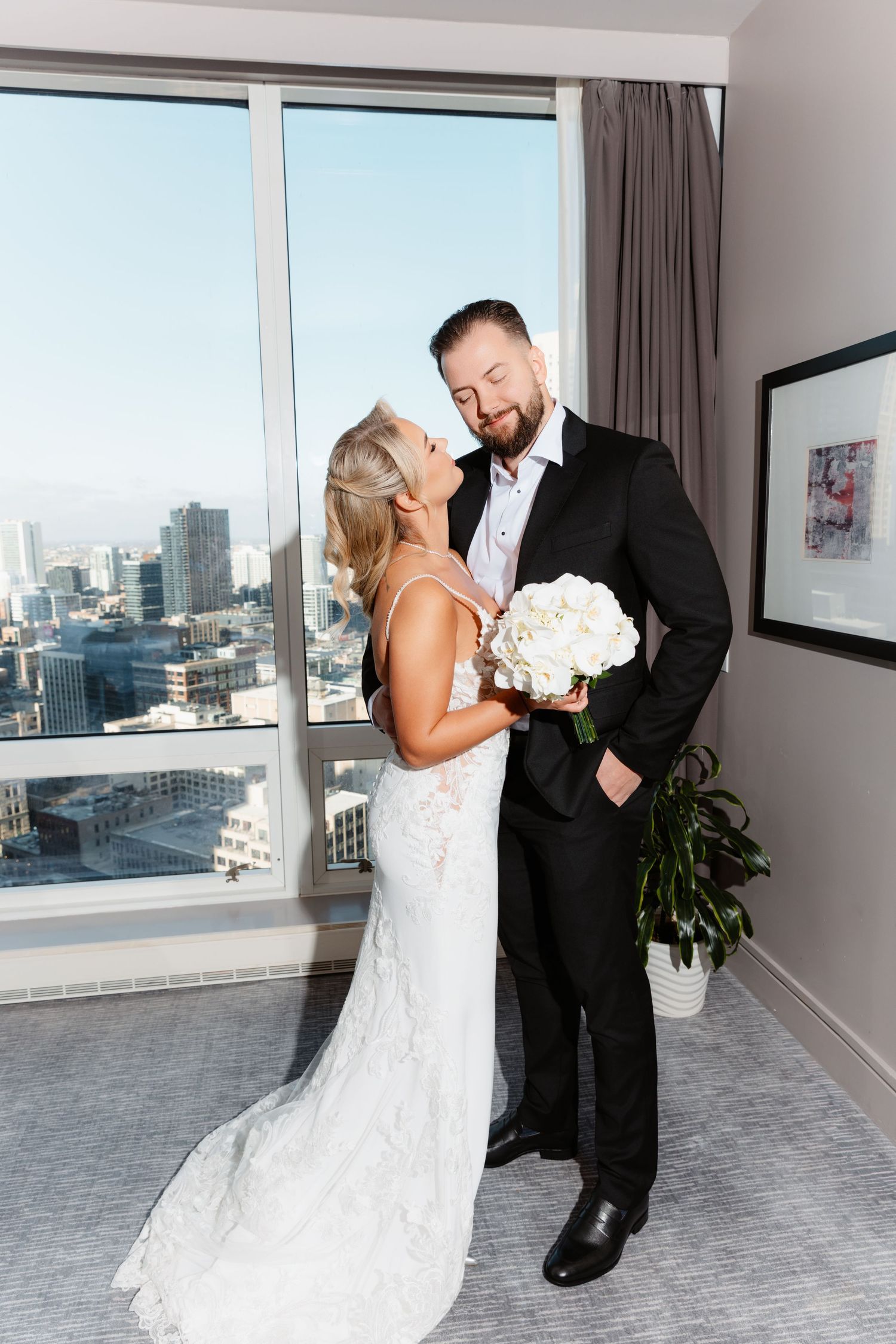 Bride in lace mermaid gown and groom in black suit share a kiss holding white bouquet in sunlit high-rise hotel room.