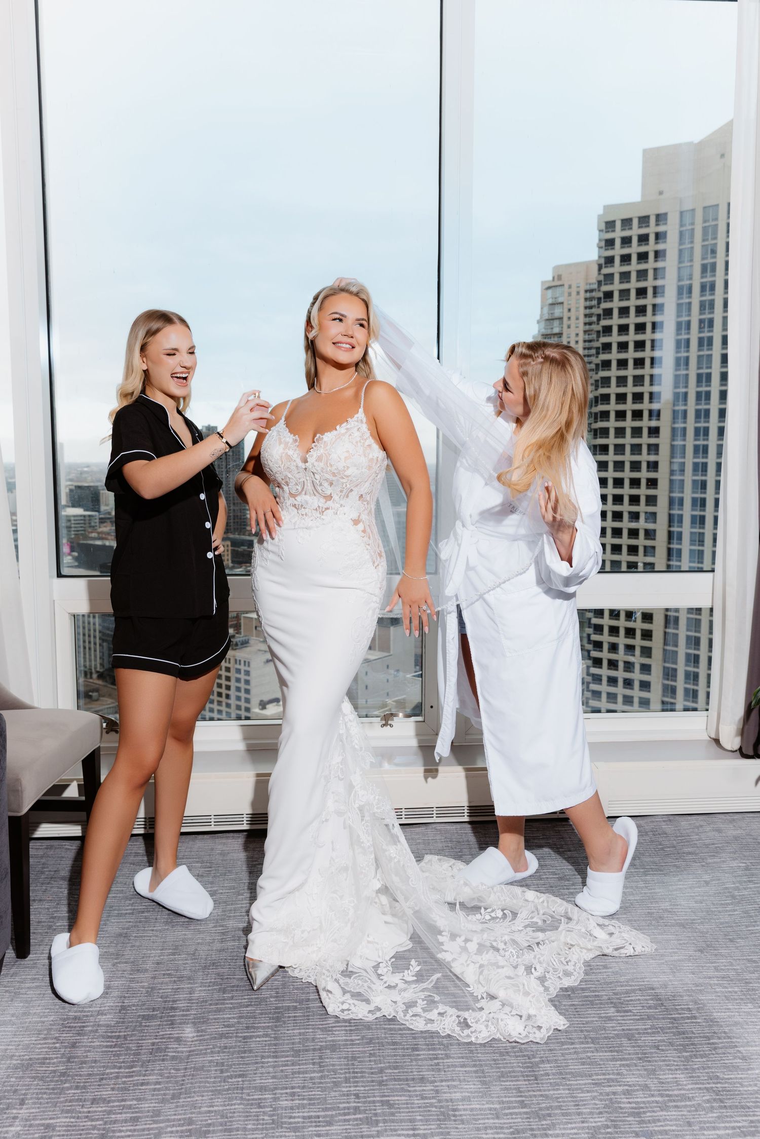 Bride in fitted lace wedding gown being helped with veil by two bridesmaids in hotel room.