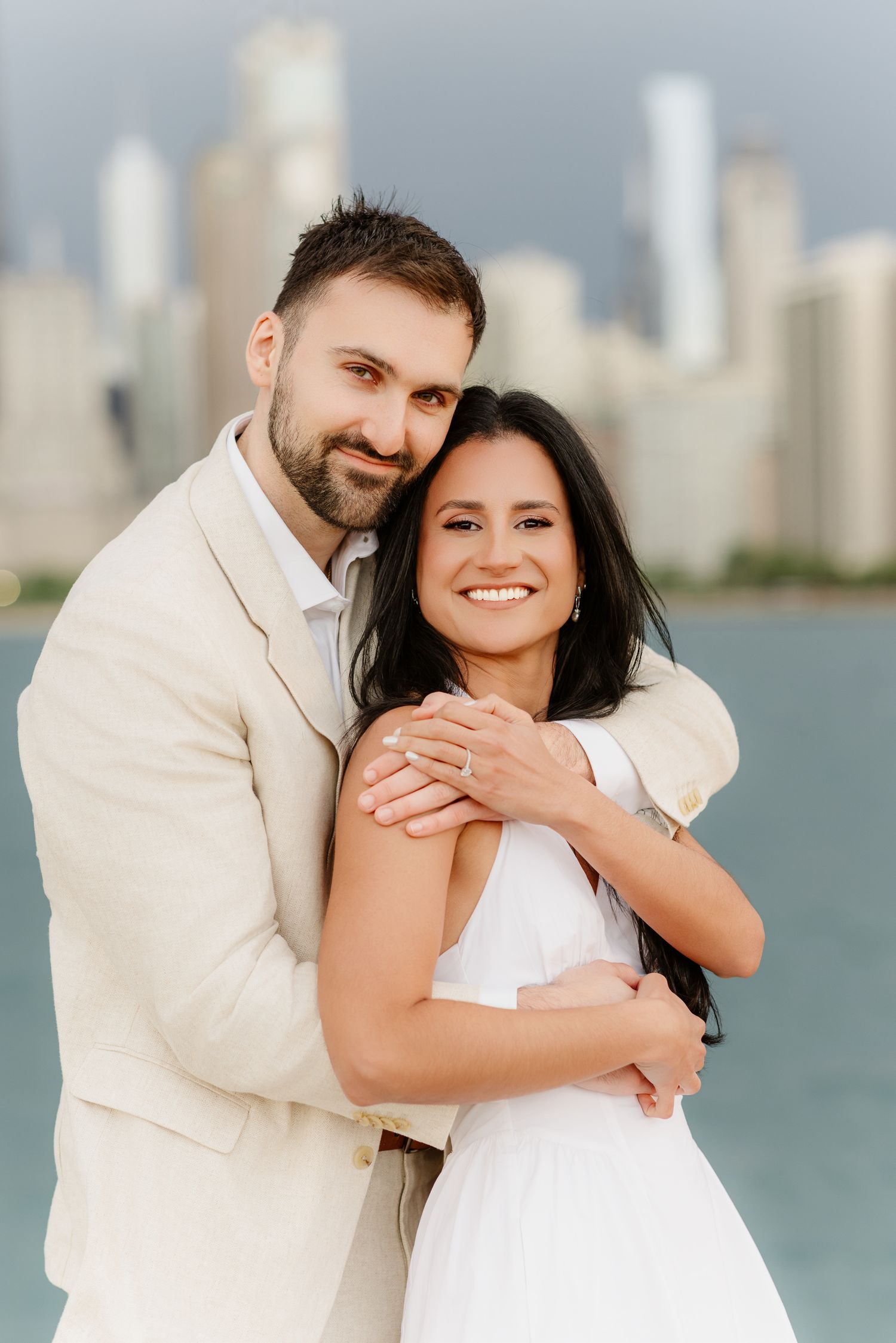 Smiling couple posing together by Chicago lakefront, man in beige suit embracing woman in white dress, skyline behind.