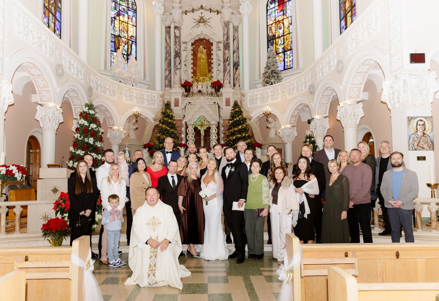 Large group wedding photo with priest kneeling in foreground inside festively decorated Catholic church at Christmas.