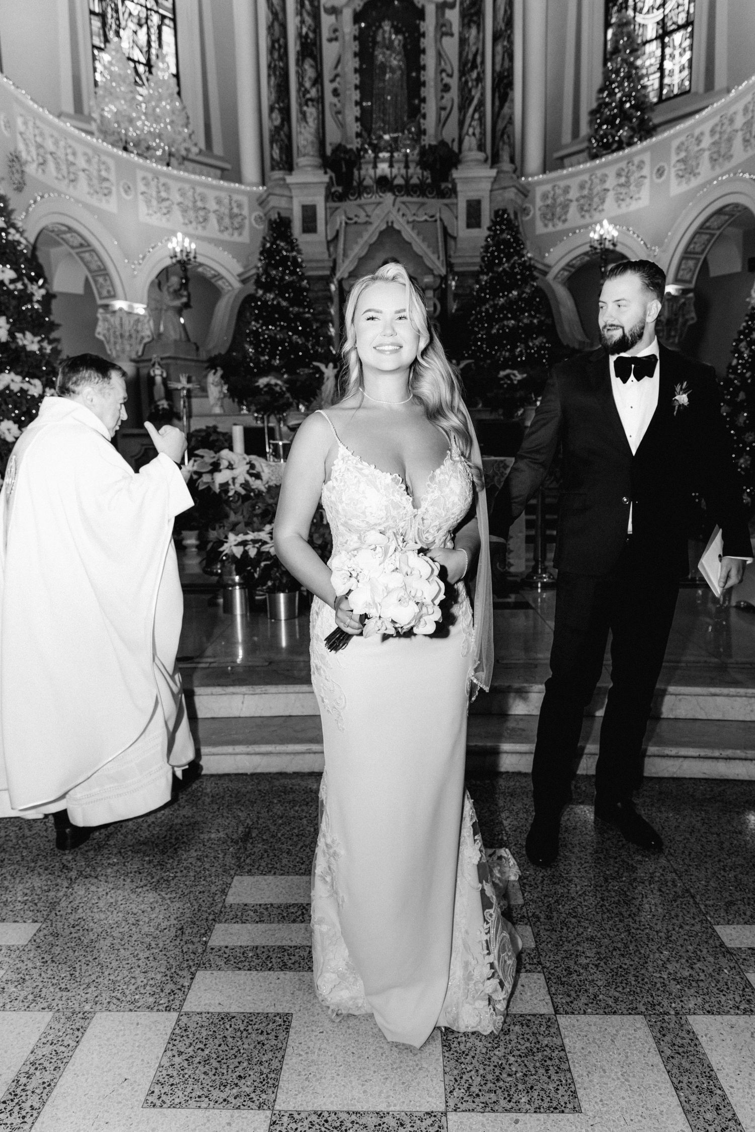 Black and white photo of bride holding bouquet at church altar with groom and priest in background.