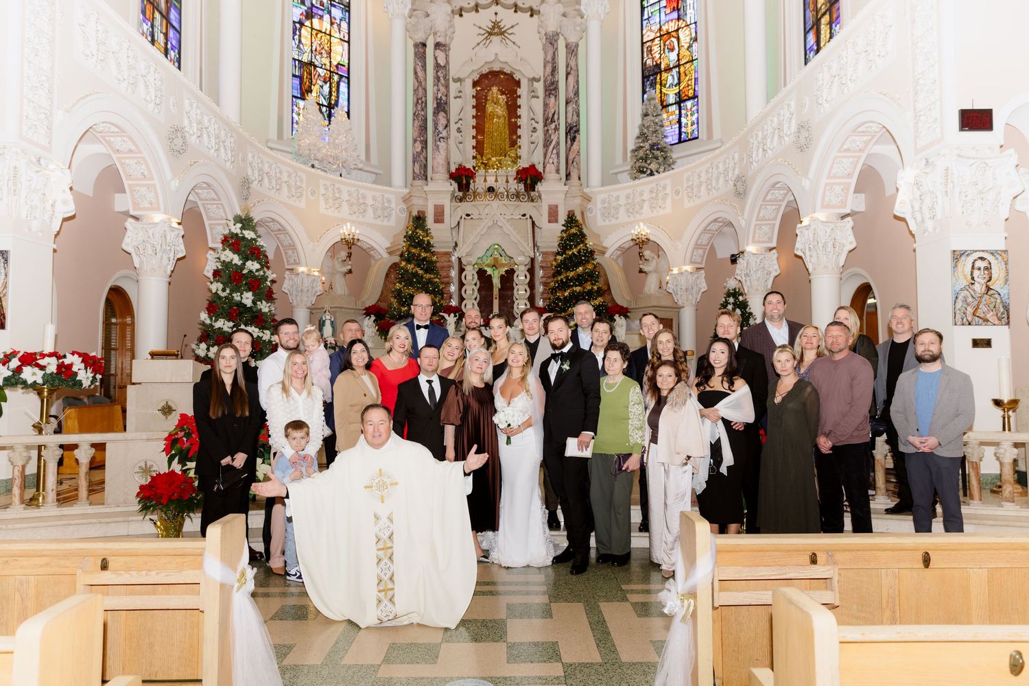 Wedding guests and bridal party pose with priest inside ornate church decorated with Christmas trees and poinsettias.