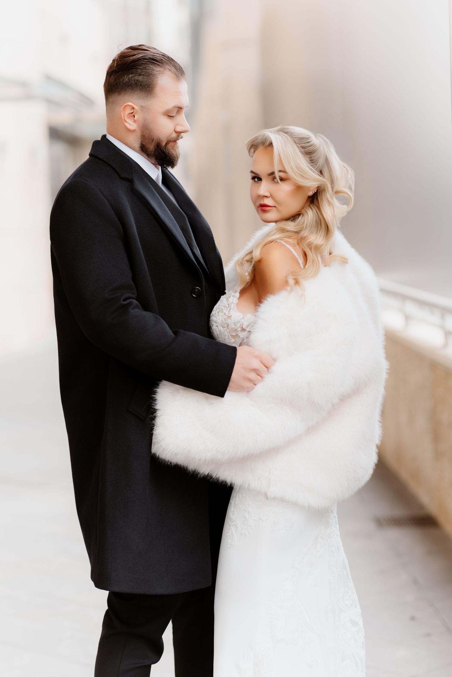 Bride in white lace gown and fur wrap embraces groom in black suit during winter wedding portrait.