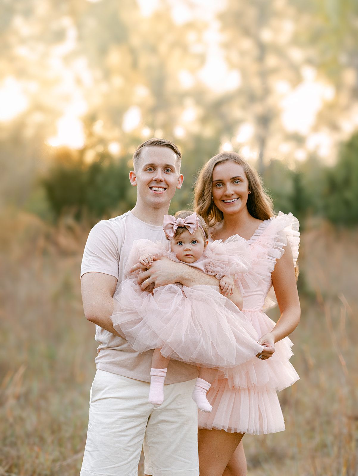 Smiling family with father holding baby in pink tutu and mother in pink dress in sunny field.