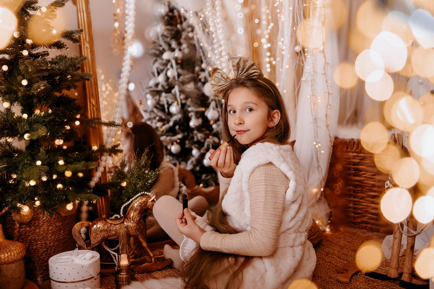 A young girl in a white fur vest sits cross-legged near a Christmas tree surrounded by golden bokeh lights.