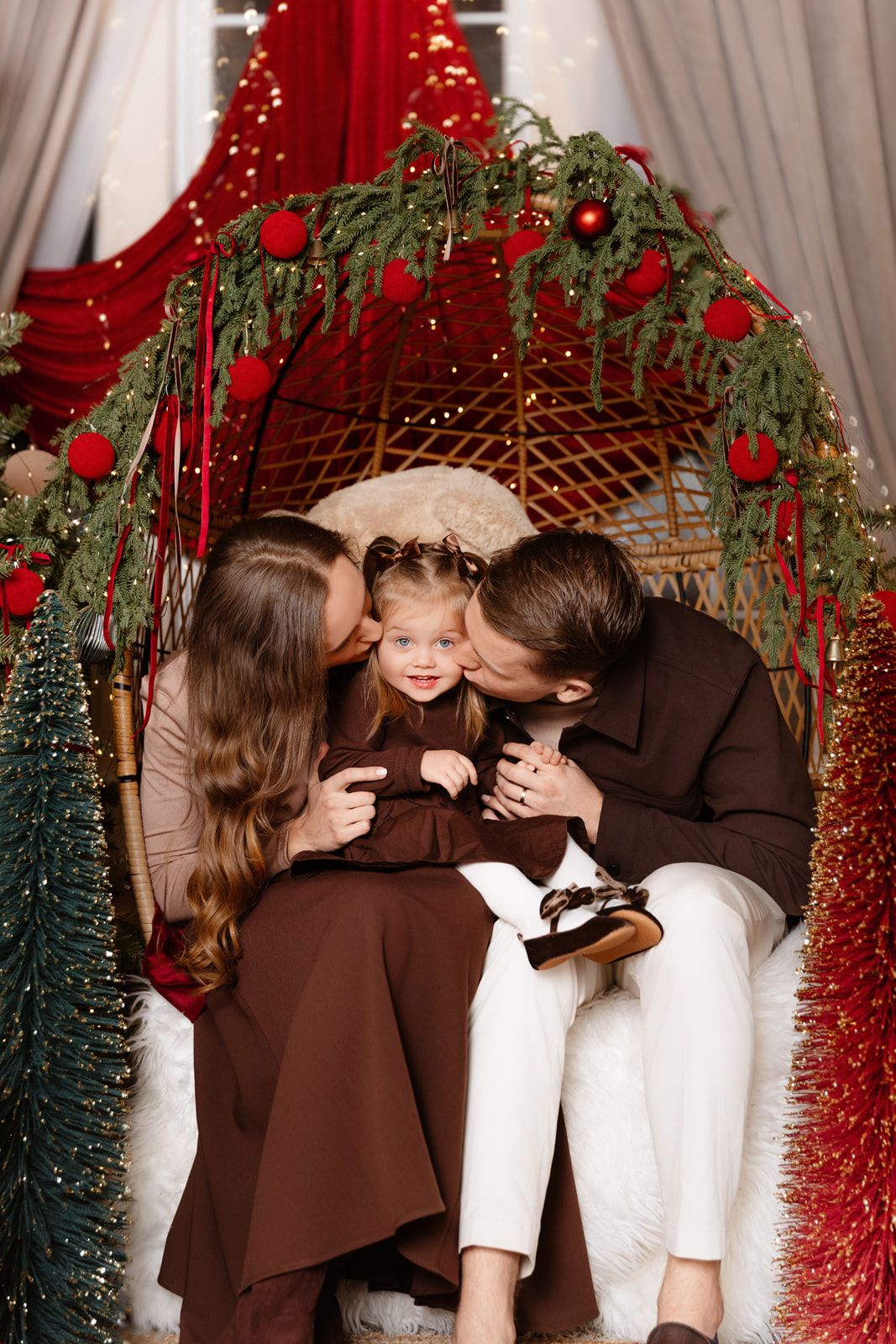 Family Christmas photo with parents kissing toddler girl in festive decorated arch with red ornaments.