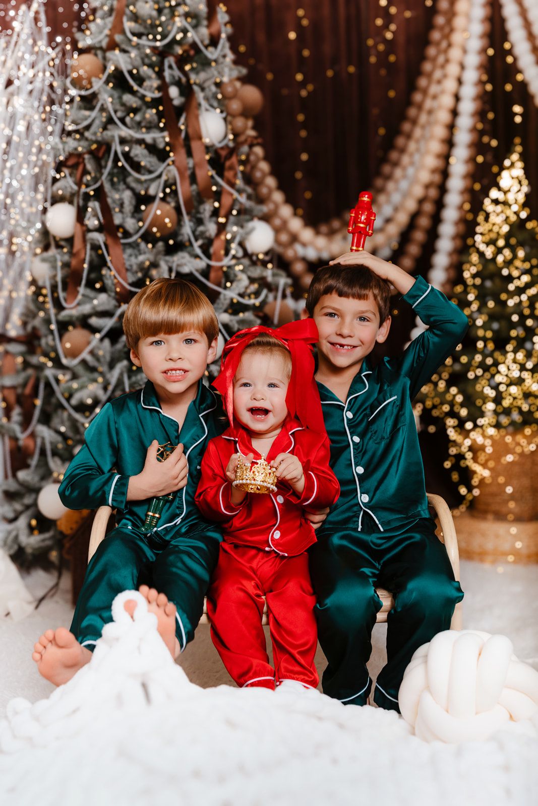 Three siblings in green and red Christmas pajamas sitting together by decorated holiday trees with lights.