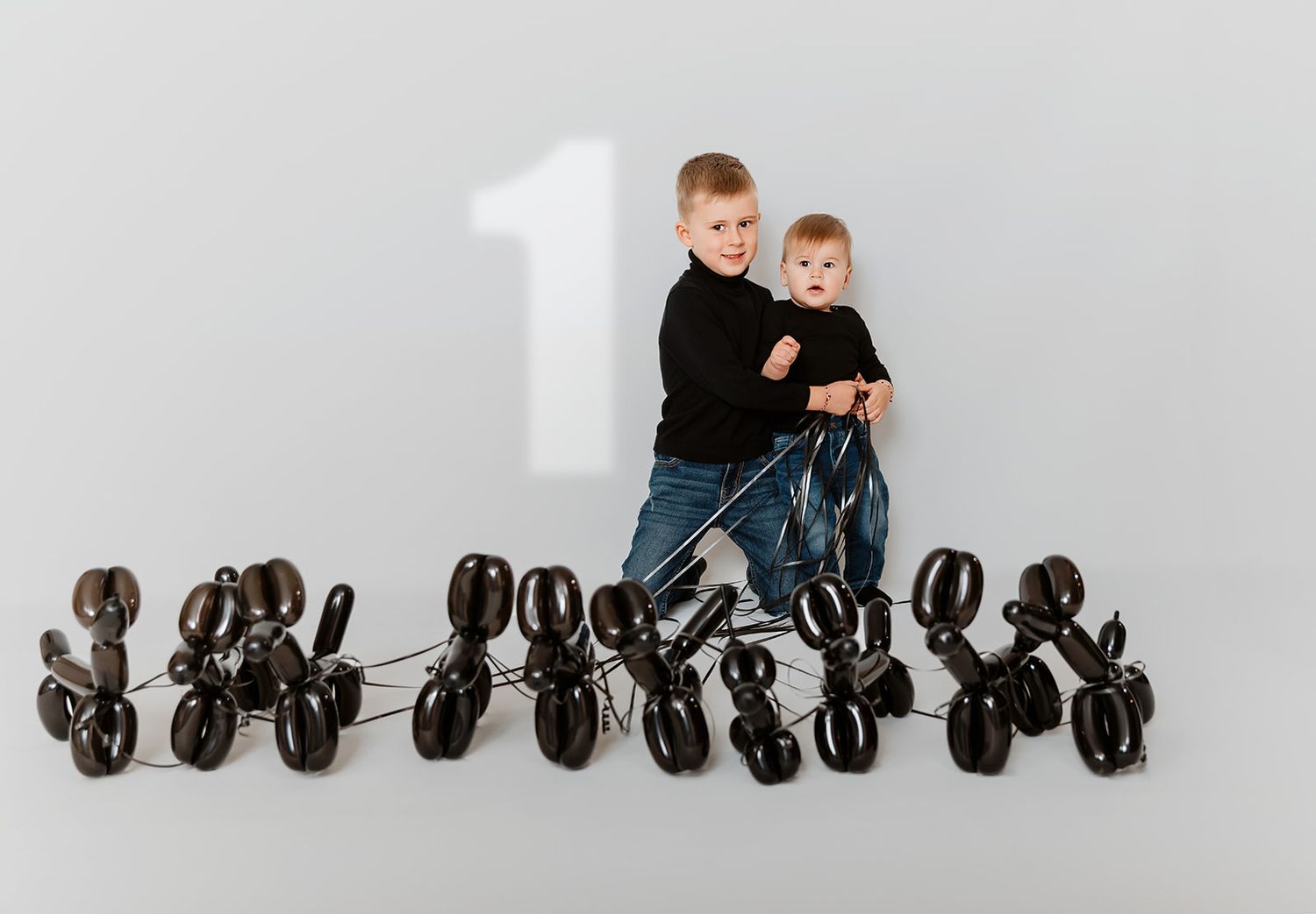 Two boys pose with black balloon animal dogs and a large number one on a gray background.