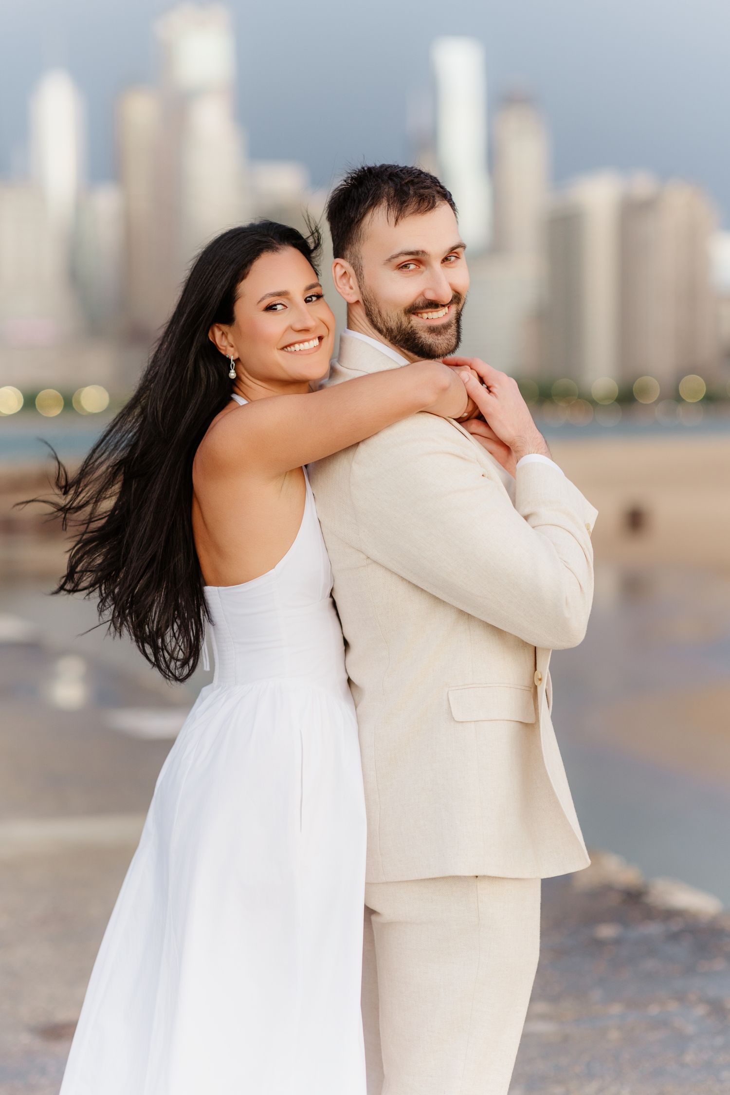 Happy couple embracing back-to-front on Chicago lakefront, both smiling with city skyline behind them.