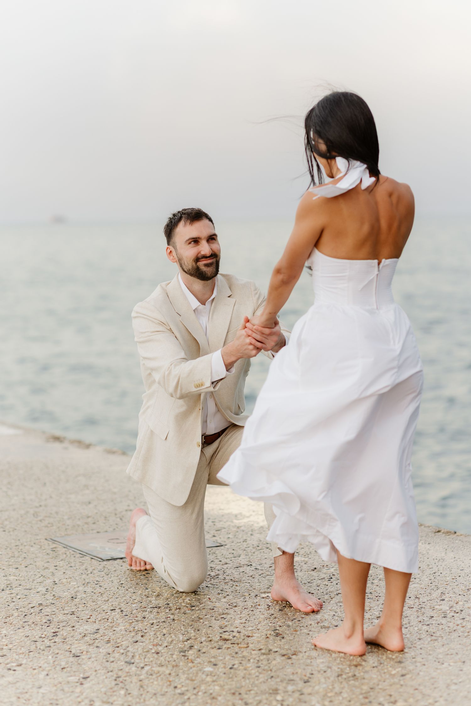 Man kneeling on waterfront path holding woman's hands during romantic seaside marriage proposal.