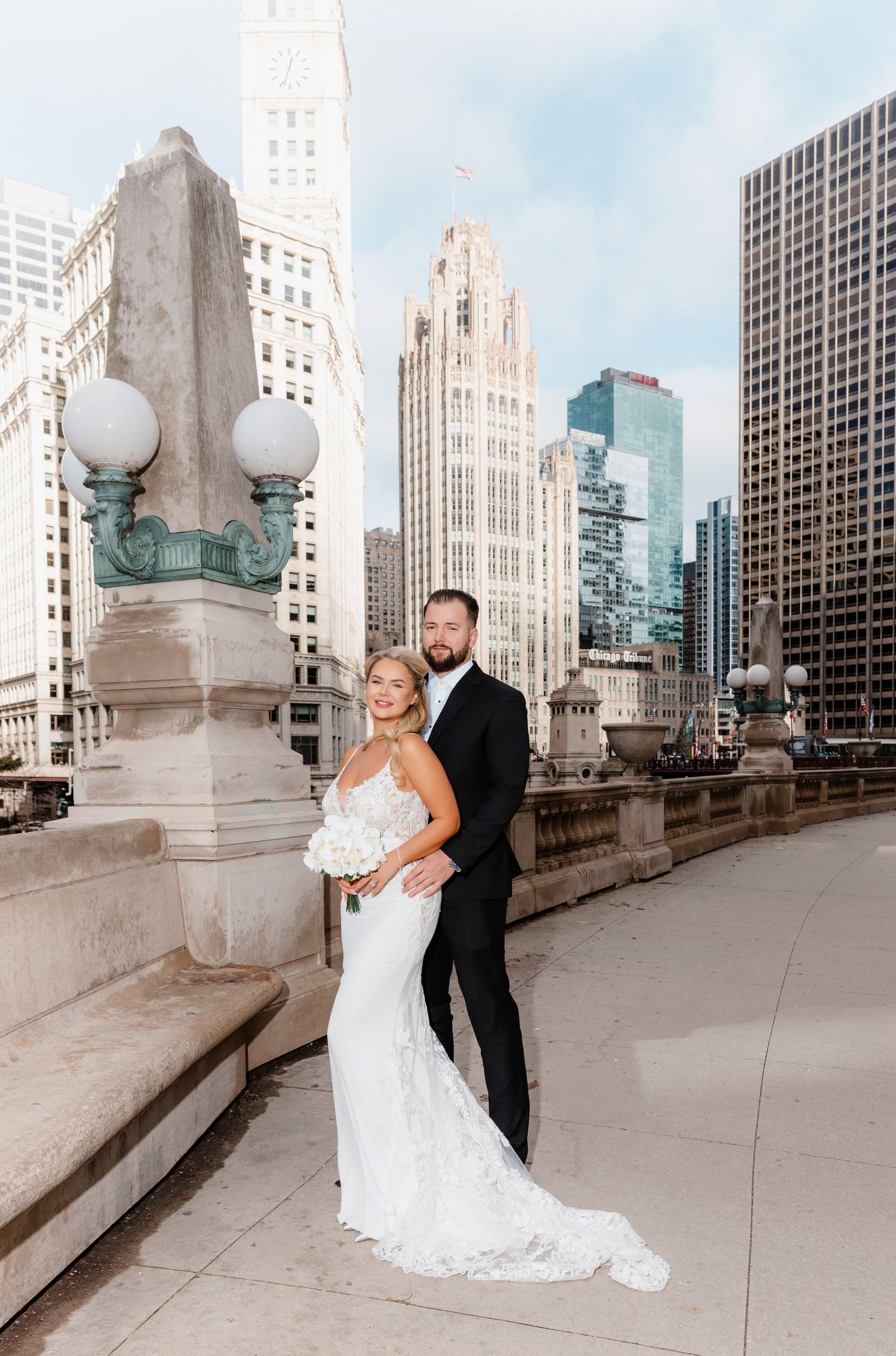 Bride in lace mermaid gown with train stands with groom, Tribune Tower visible in Chicago skyline.