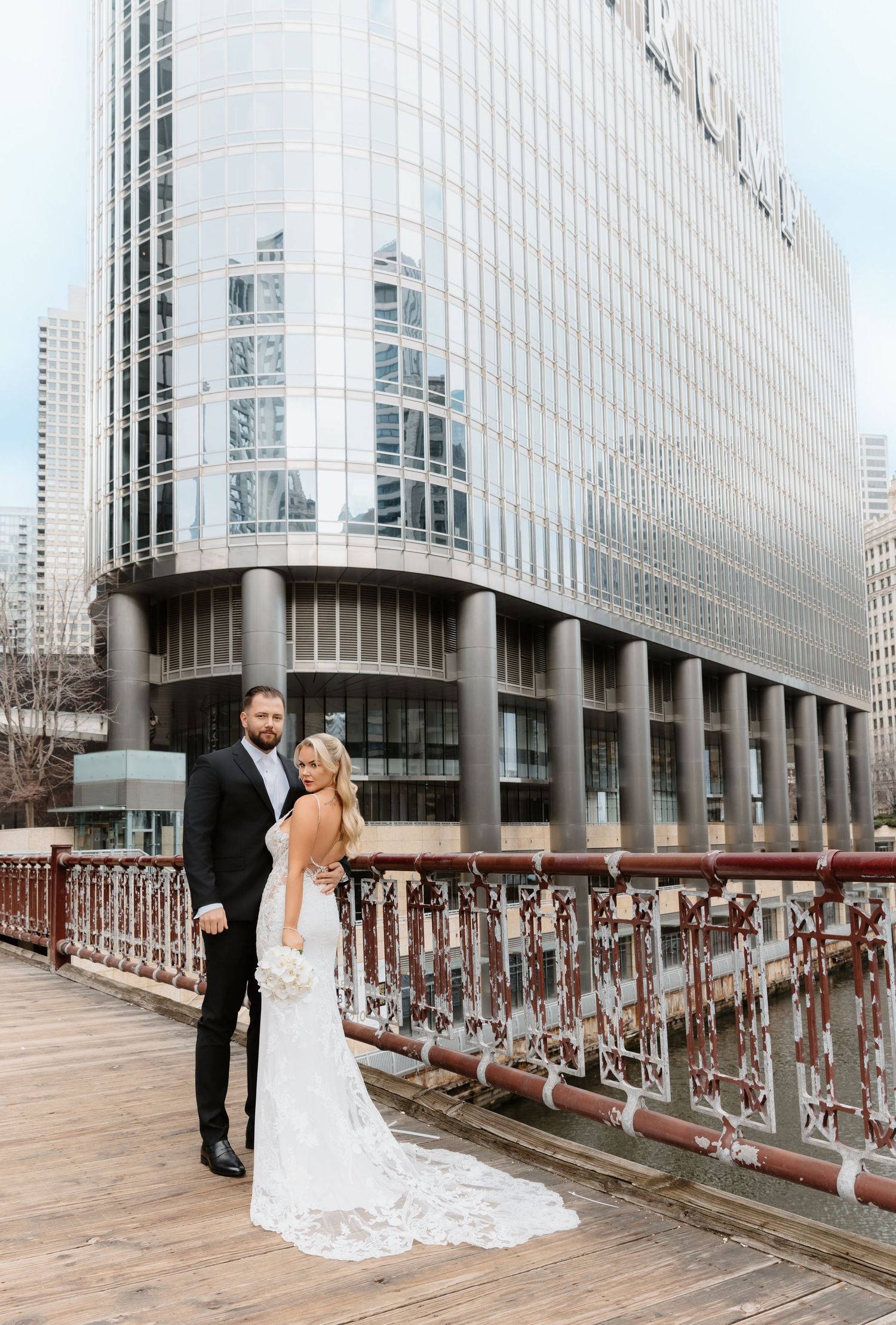 Bride in backless lace gown and groom in tuxedo pose on Chicago riverwalk with glass skyscraper backdrop.