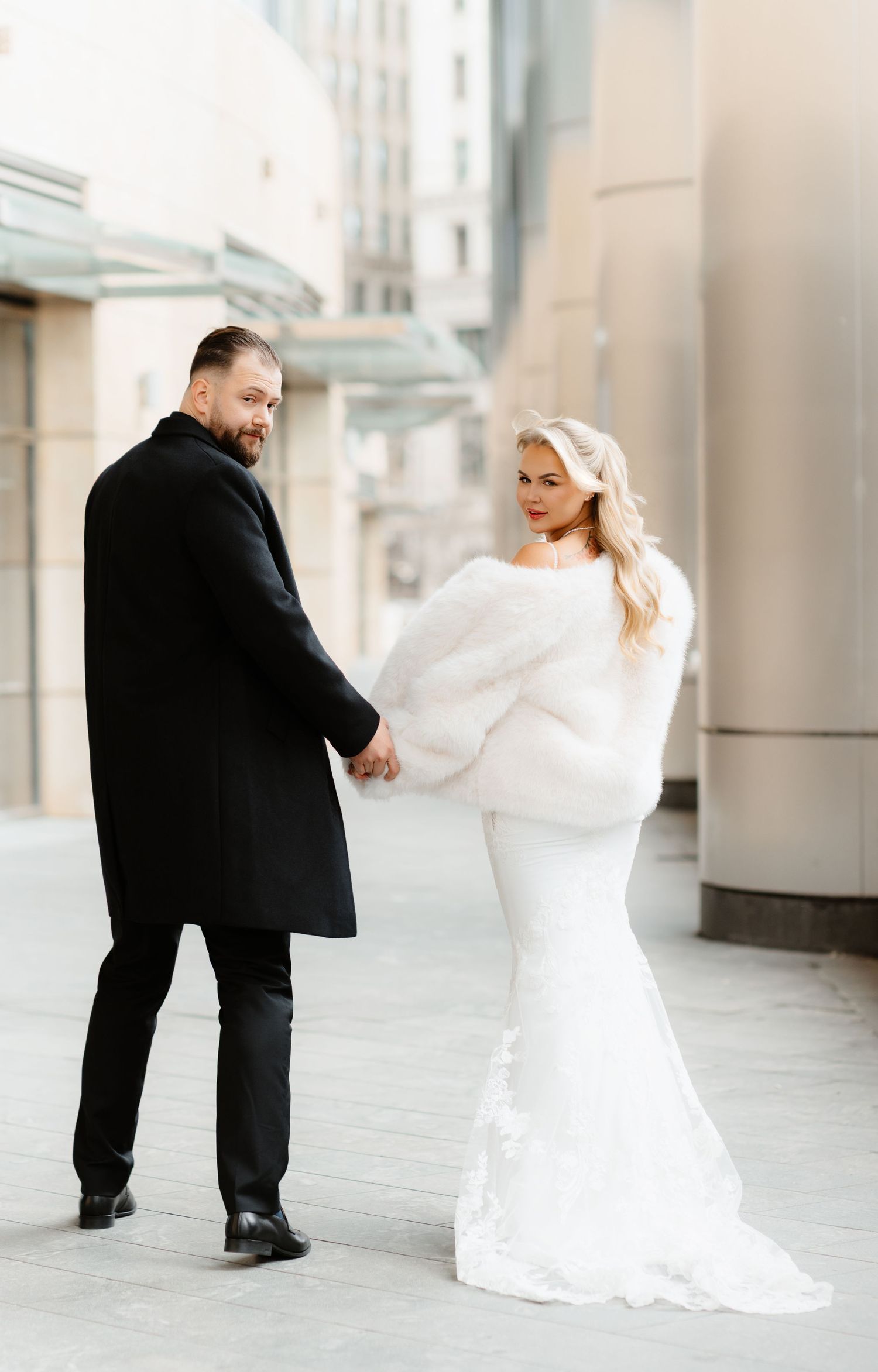 Bride in fur stole and groom in black coat hold hands while looking back on city street.
