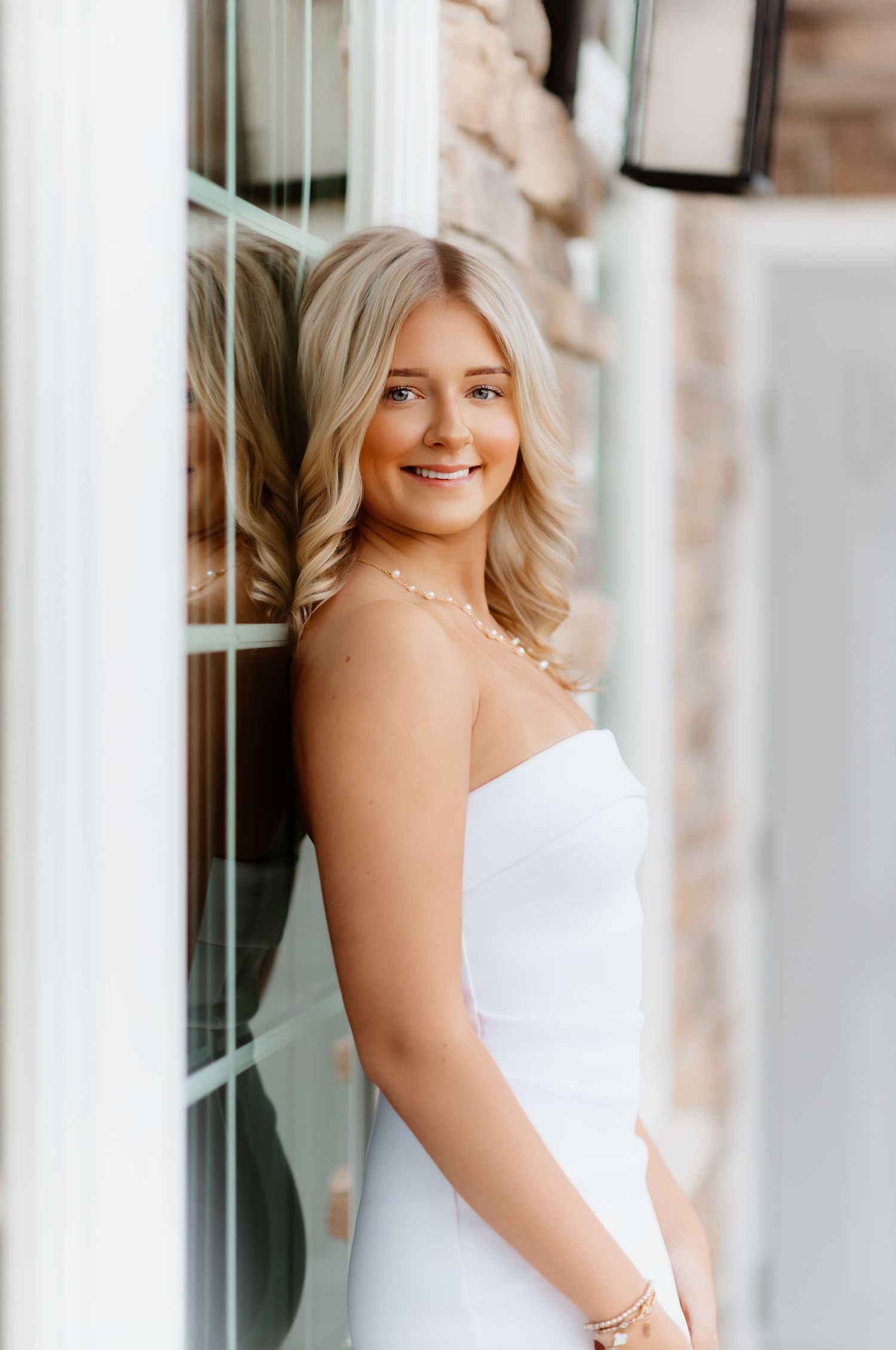 Smiling blonde woman in white strapless dress leaning against window with reflection.