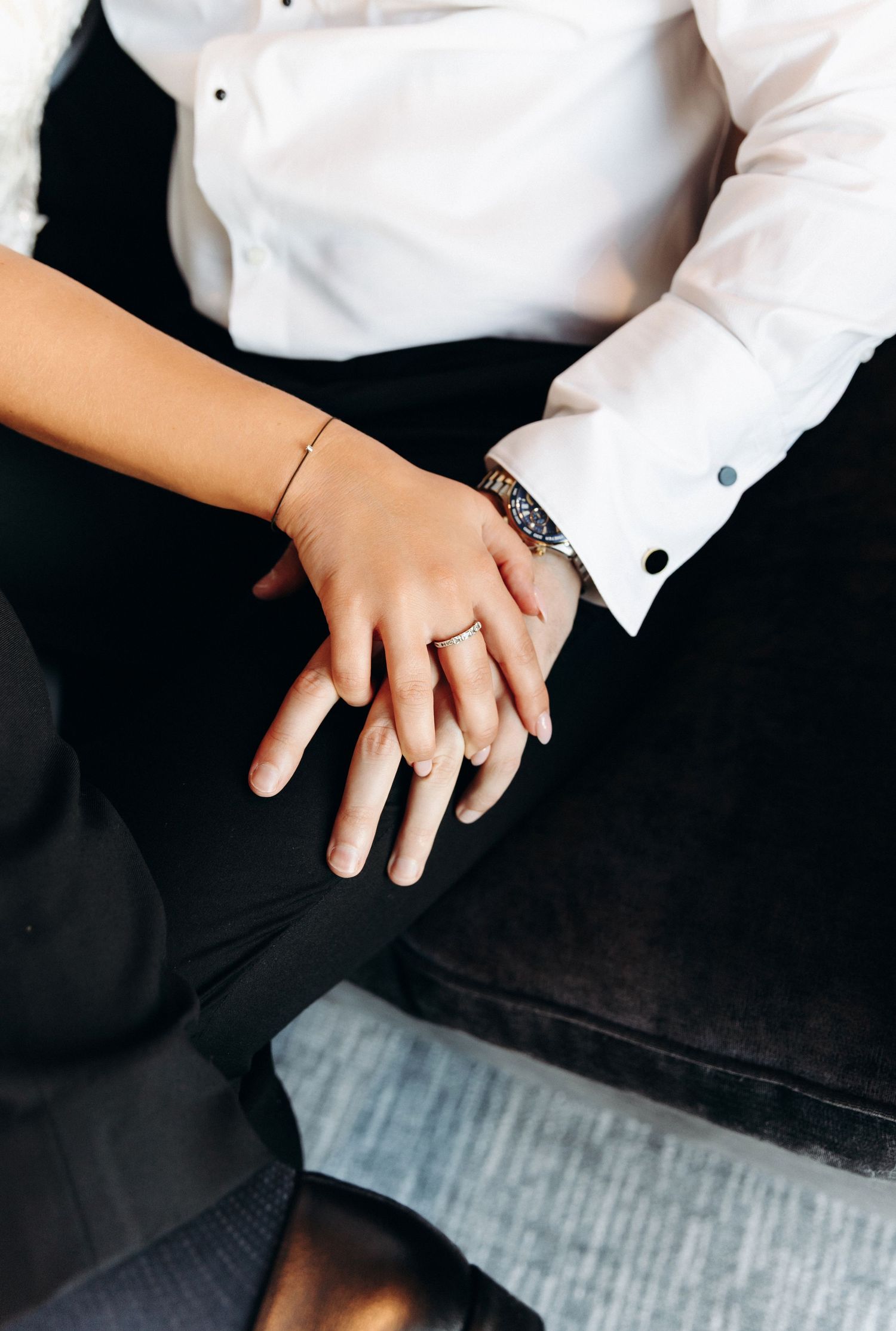 Bride and groom holding hands showing wedding rings and gold bracelet while seated together.