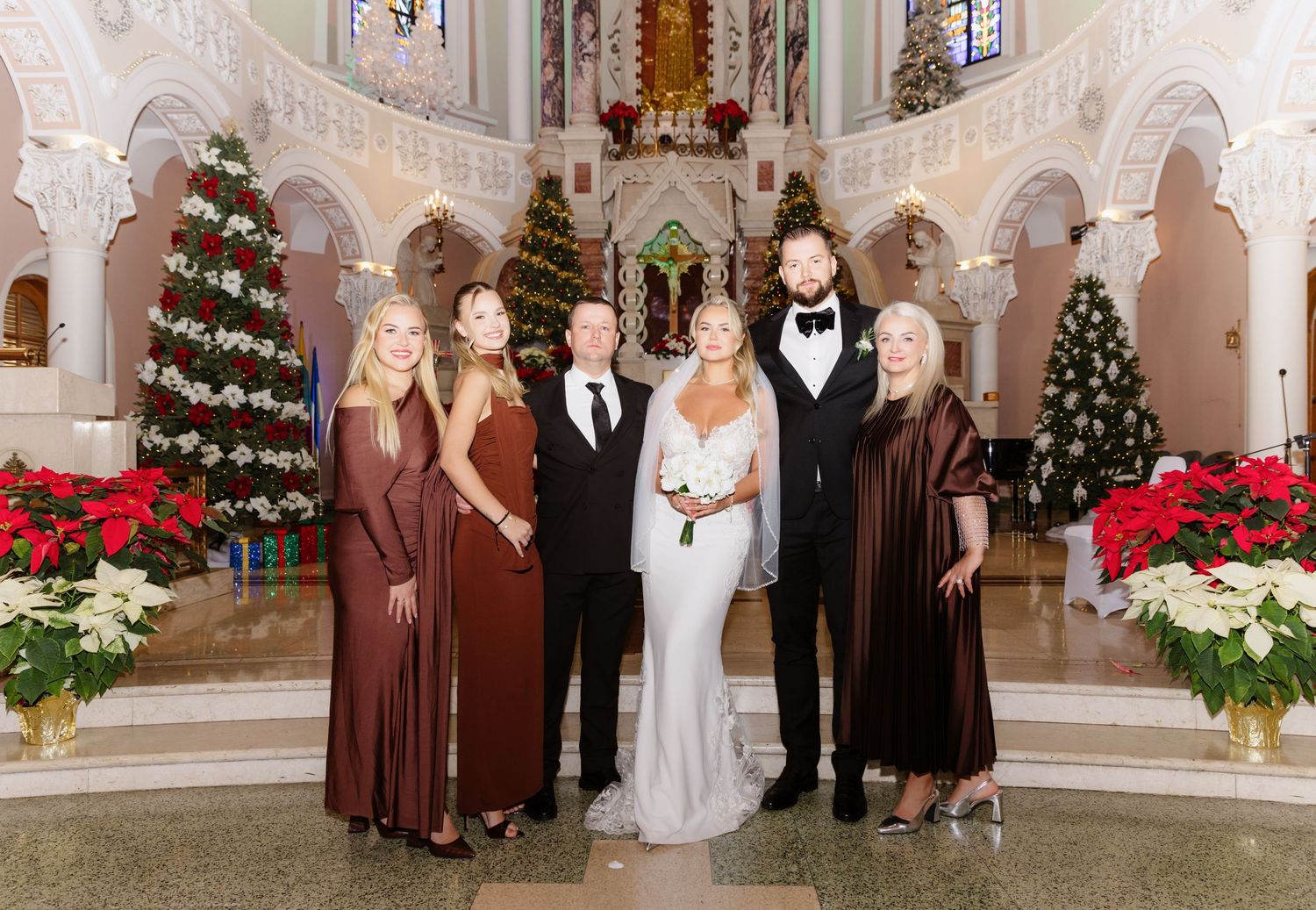 Wedding party poses in church with Christmas decorations, bride in white gown flanked by bridesmaids in burgundy dresses.