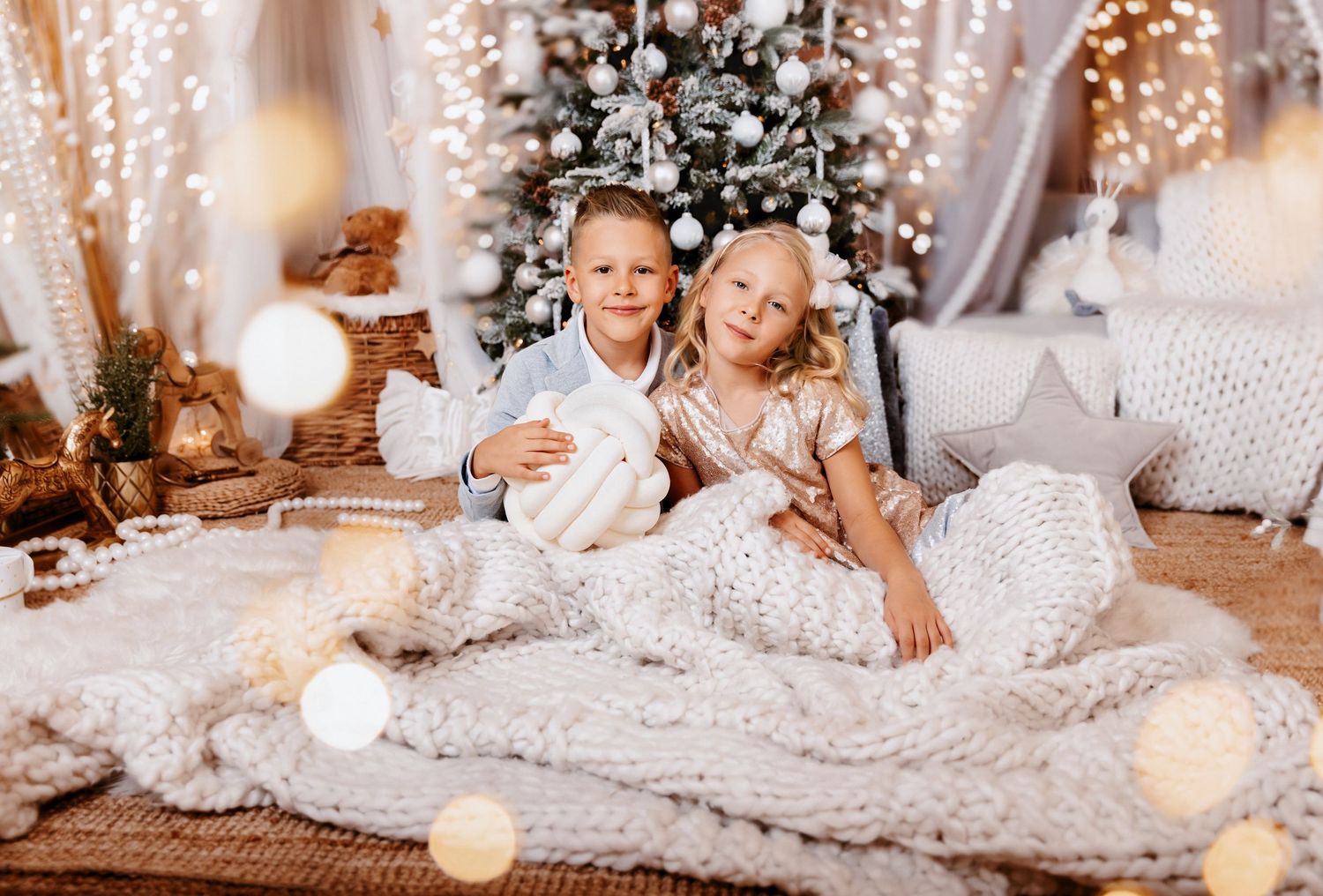 A boy and girl cuddle together under a white knit blanket in front of a snow-dusted Christmas tree with warm lights.