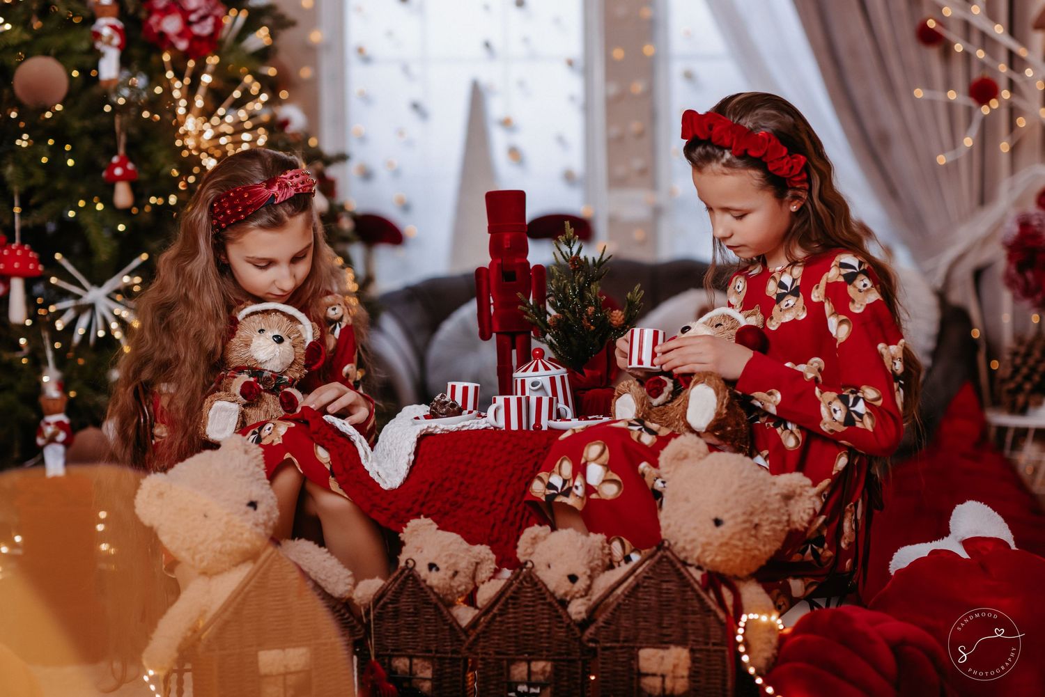 Two girls in red floral pajamas play with teddy bears and wicker houses beside a glowing Christmas tree.