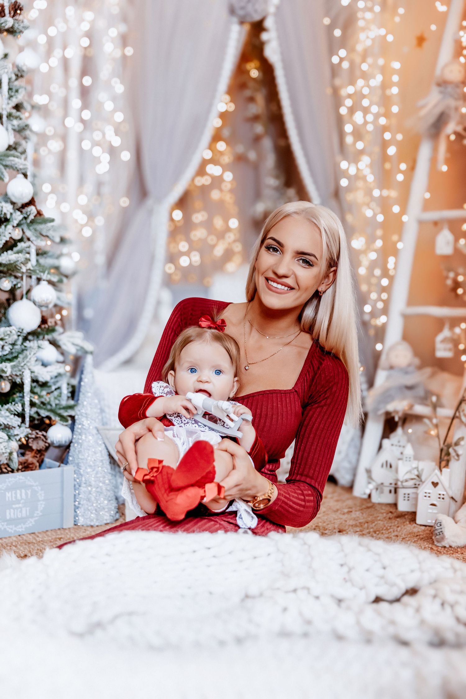 Smiling blonde woman in red holds baby girl in Christmas decorated room with warm bokeh lights.