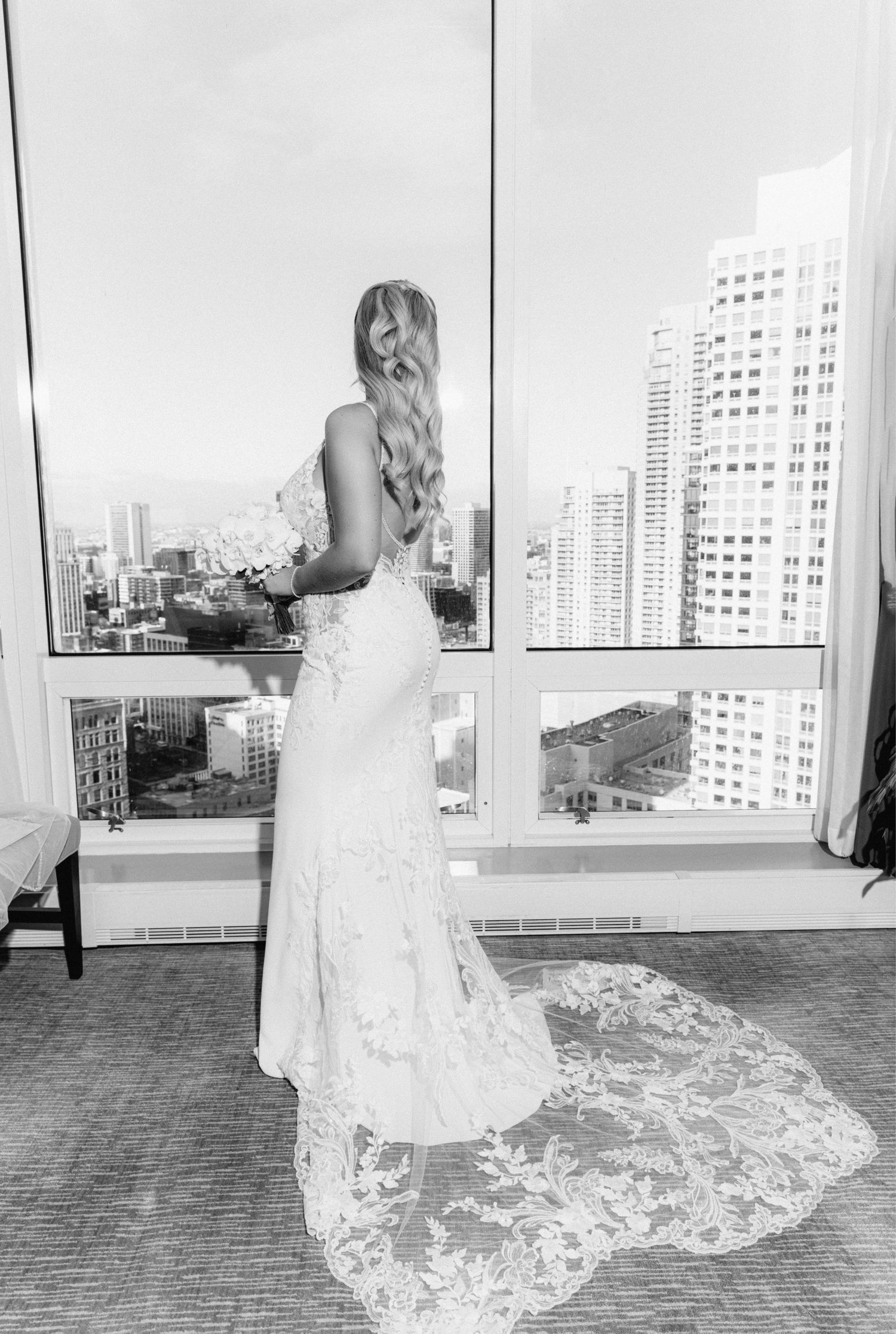 Black and white photo of bride in lace wedding gown with train gazing at city skyline.