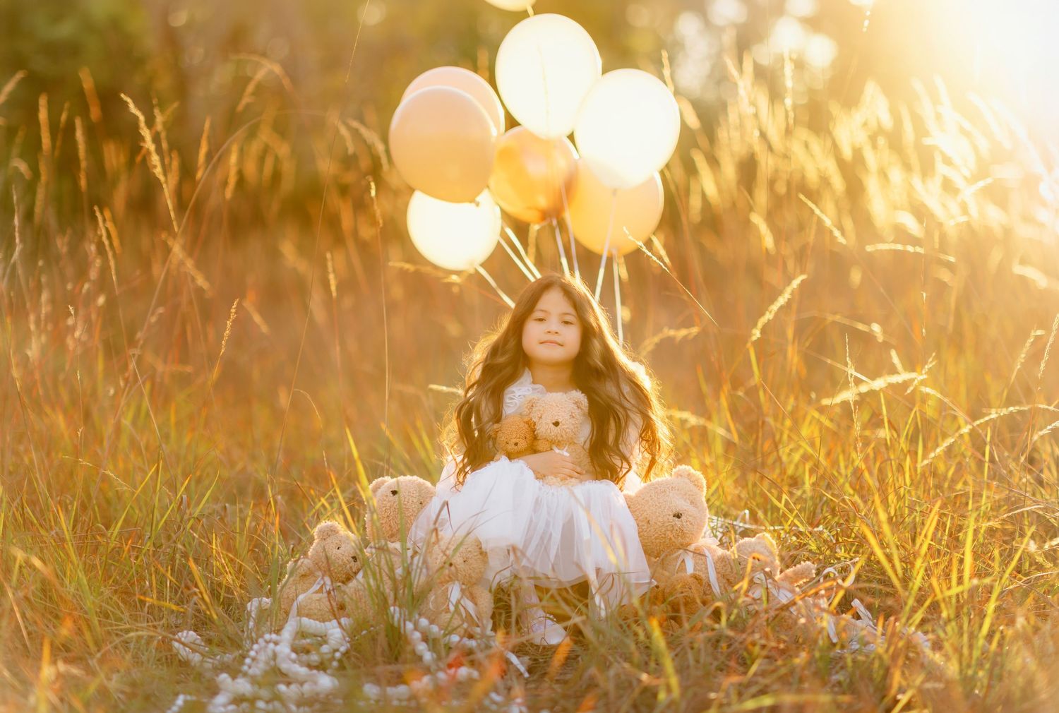 Young girl in white dress sits in golden field holding teddy bears with yellow balloons in warm sunlight.