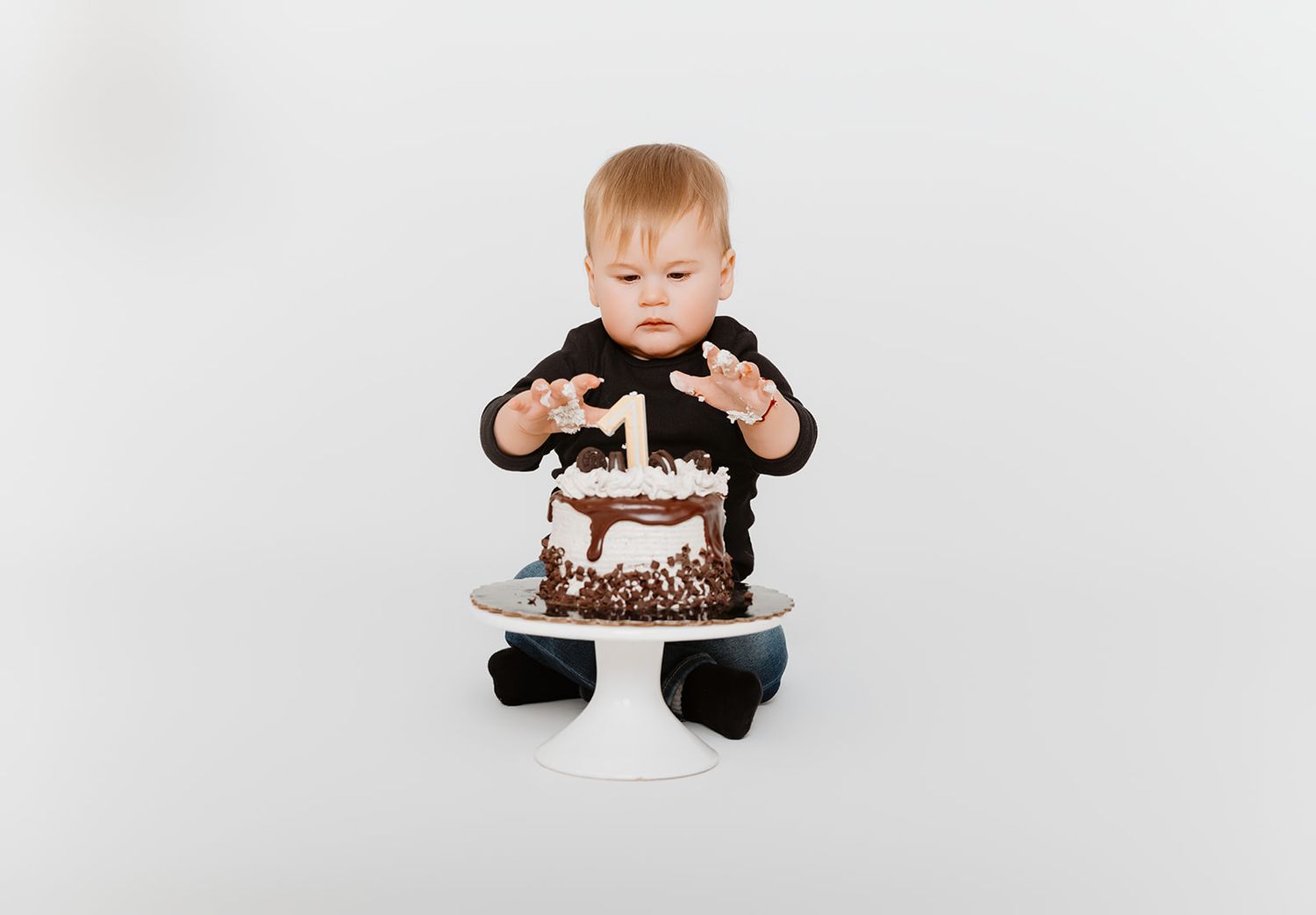Baby in black outfit reaches toward a chocolate first birthday cake on a white cake stand.